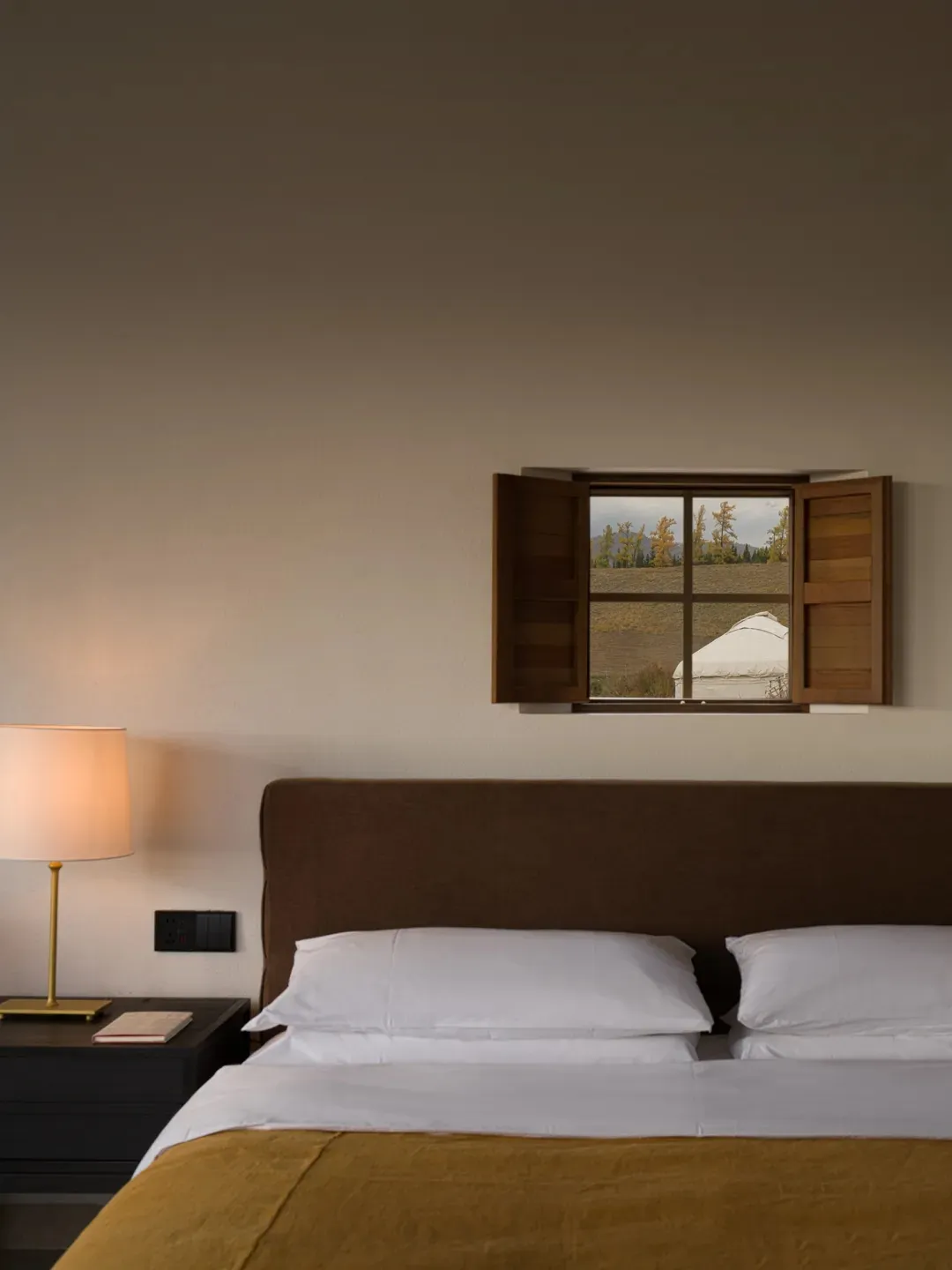 Modern minimalist bedroom with brown upholstered headboard, white bedding, mustard yellow linen throw, brass table lamp, and open walnut wooden shutter window revealing a view of autumn trees and a white yurt.