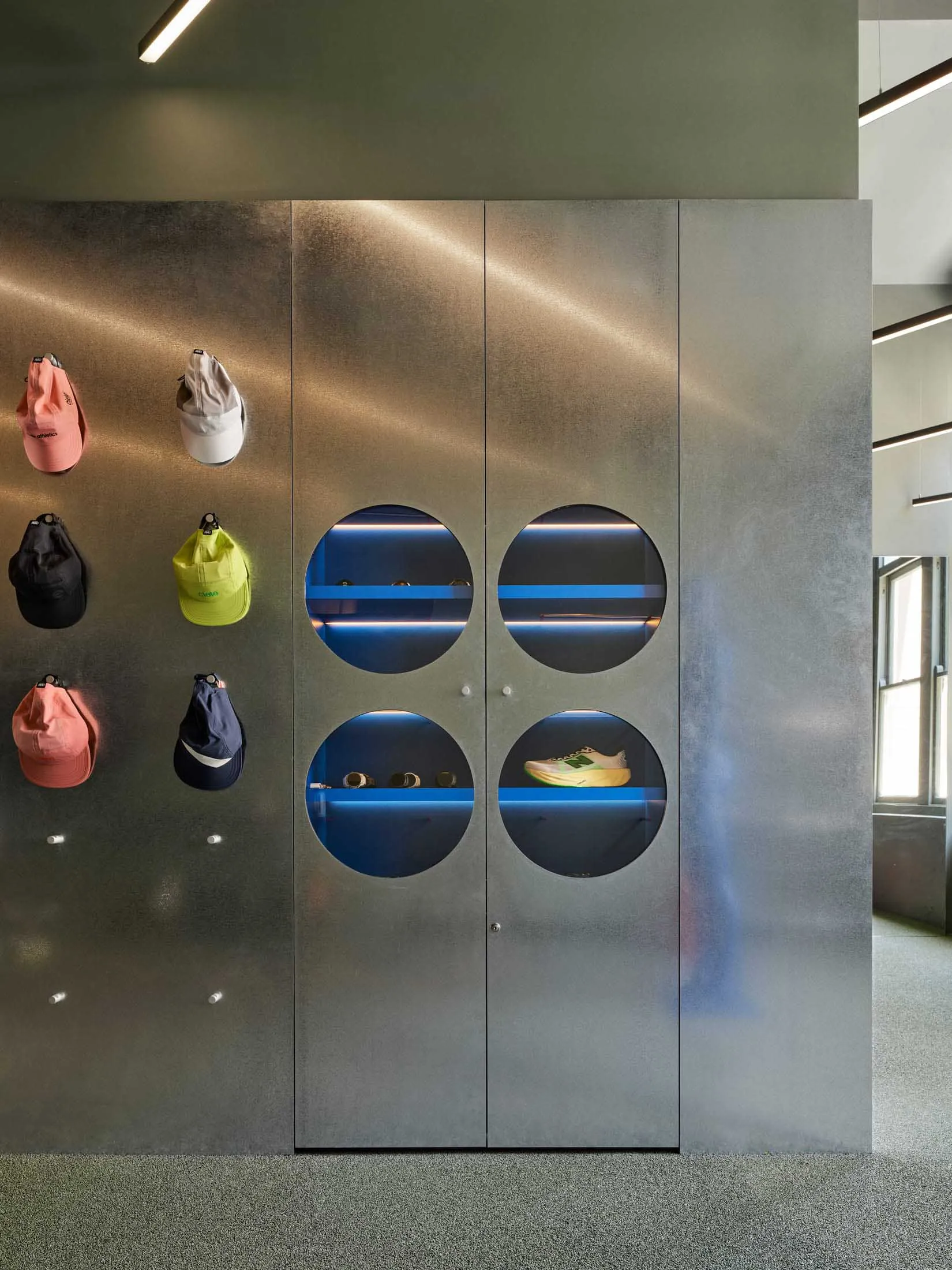 Modern retail wall featuring brushed stainless steel cabinetry with four circular illuminated display niches containing sneakers and accessories, accompanied by colorful baseball caps hanging on the left side.
