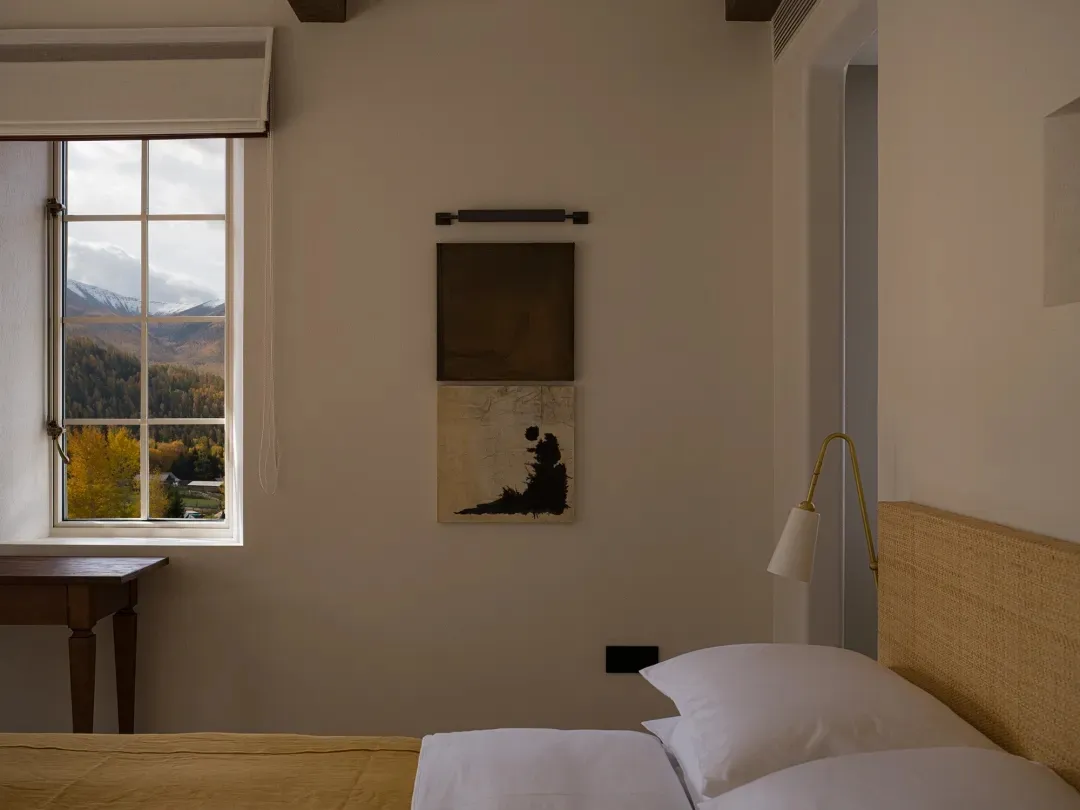 Cozy bedroom interior featuring a woven rattan headboard, white linens, mustard yellow throw, two pieces of abstract wall art, and a large window overlooking snow-capped mountains and autumn forest.