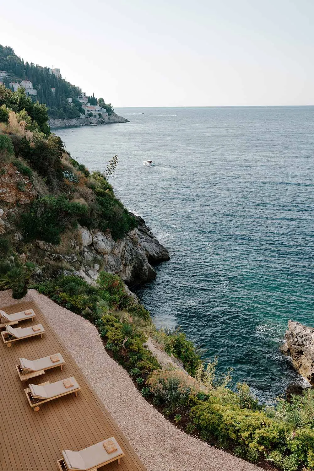 Luxurious wooden terrace with teak lounge chairs overlooking a rocky Mediterranean coastline and deep blue sea.