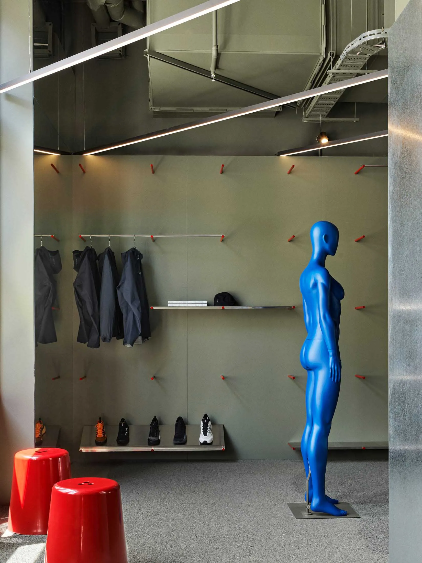 Modern athletic locker room with sage green walls, stainless steel benches and hooks, hanging dark sportswear, a row of sneakers, two glossy red stools, and a full-body glossy blue mannequin standing on the right.