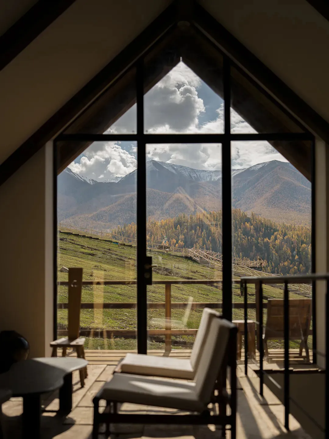 Interior view of a modern mountain lounge featuring a large black-framed gable window overlooking a vast mountain range with autumn foliage and snow-capped peaks, with minimalist wooden furniture in the foreground.