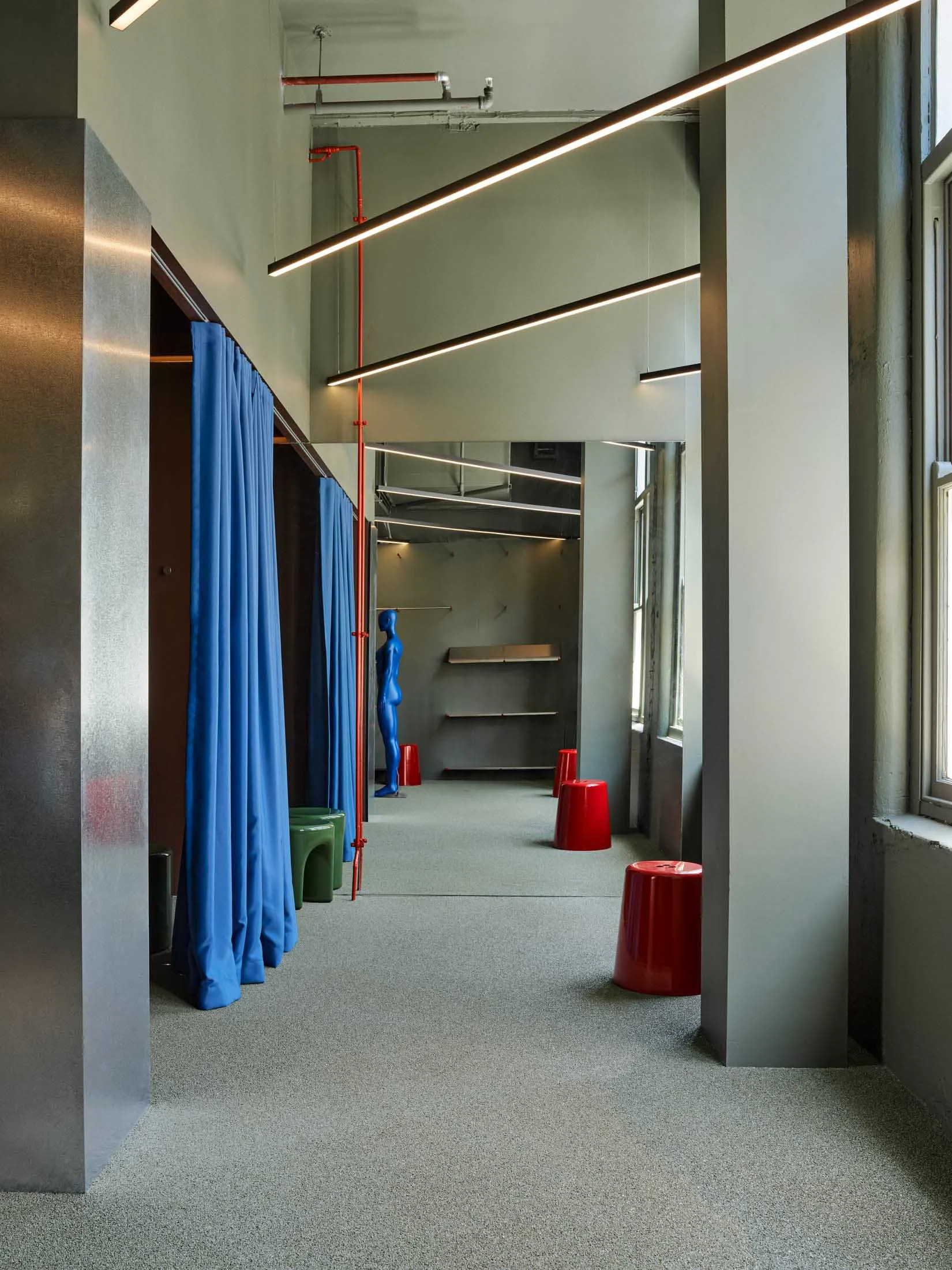 Modern industrial-style retail changing room featuring bright blue curtains, exposed red pipes, concrete walls, grey carpet, and colorful plastic stools with a blue mannequin.
