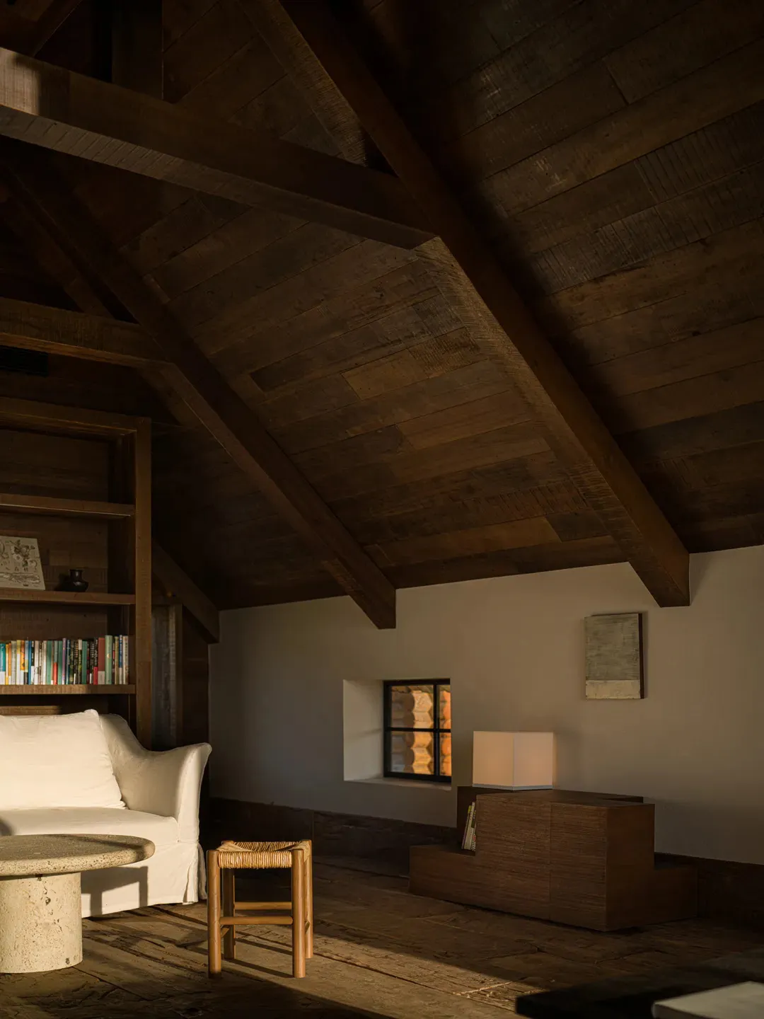 Cozy attic living room featuring reclaimed wood cathedral ceiling, white linen sofa, stone coffee table, and wooden stool in warm natural light.