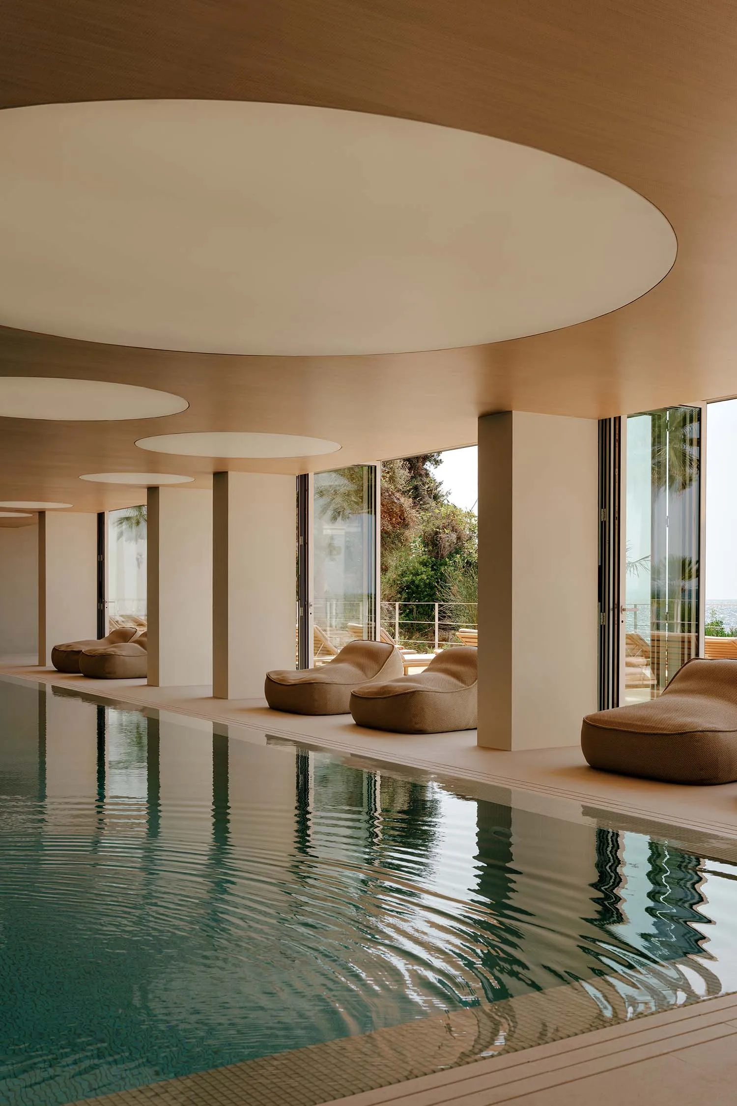 Modern indoor swimming pool with reflective turquoise water, beige fabric lounge chairs, massive circular wooden ceiling details, and large glass doors opening to a sea view.
