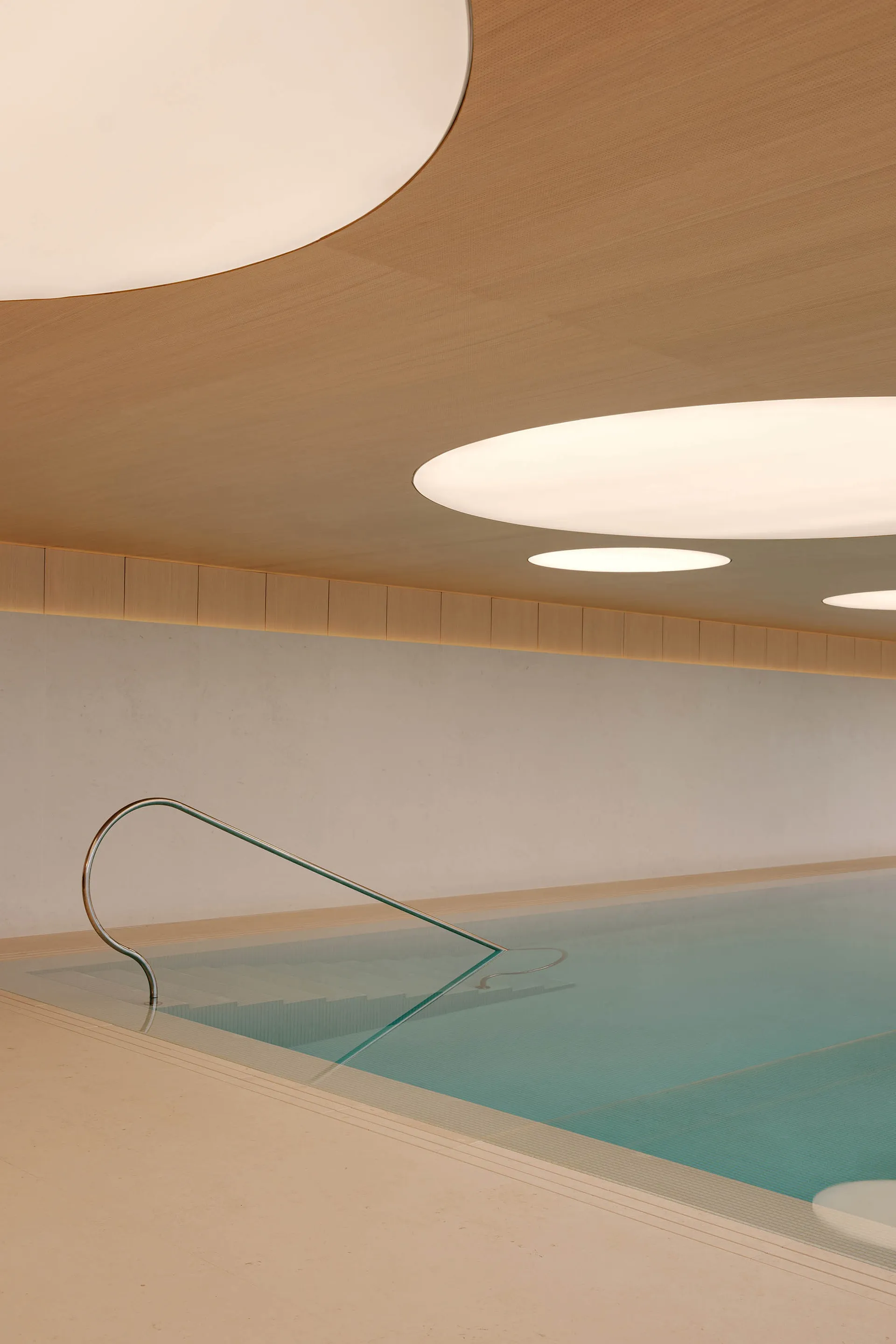 Modern indoor swimming pool with light blue water, stainless steel handrail, concrete walls and a curved wooden ceiling featuring large circular light fixtures.
