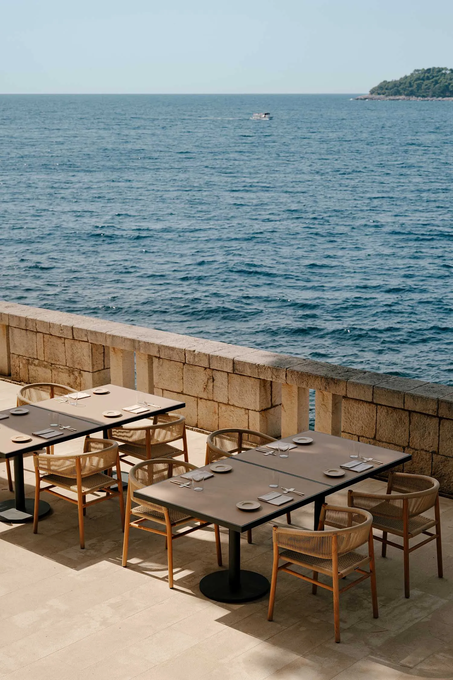 Outdoor dining tables and woven rattan chairs arranged on a stone terrace overlooking the calm blue sea with a distant boat and green island.