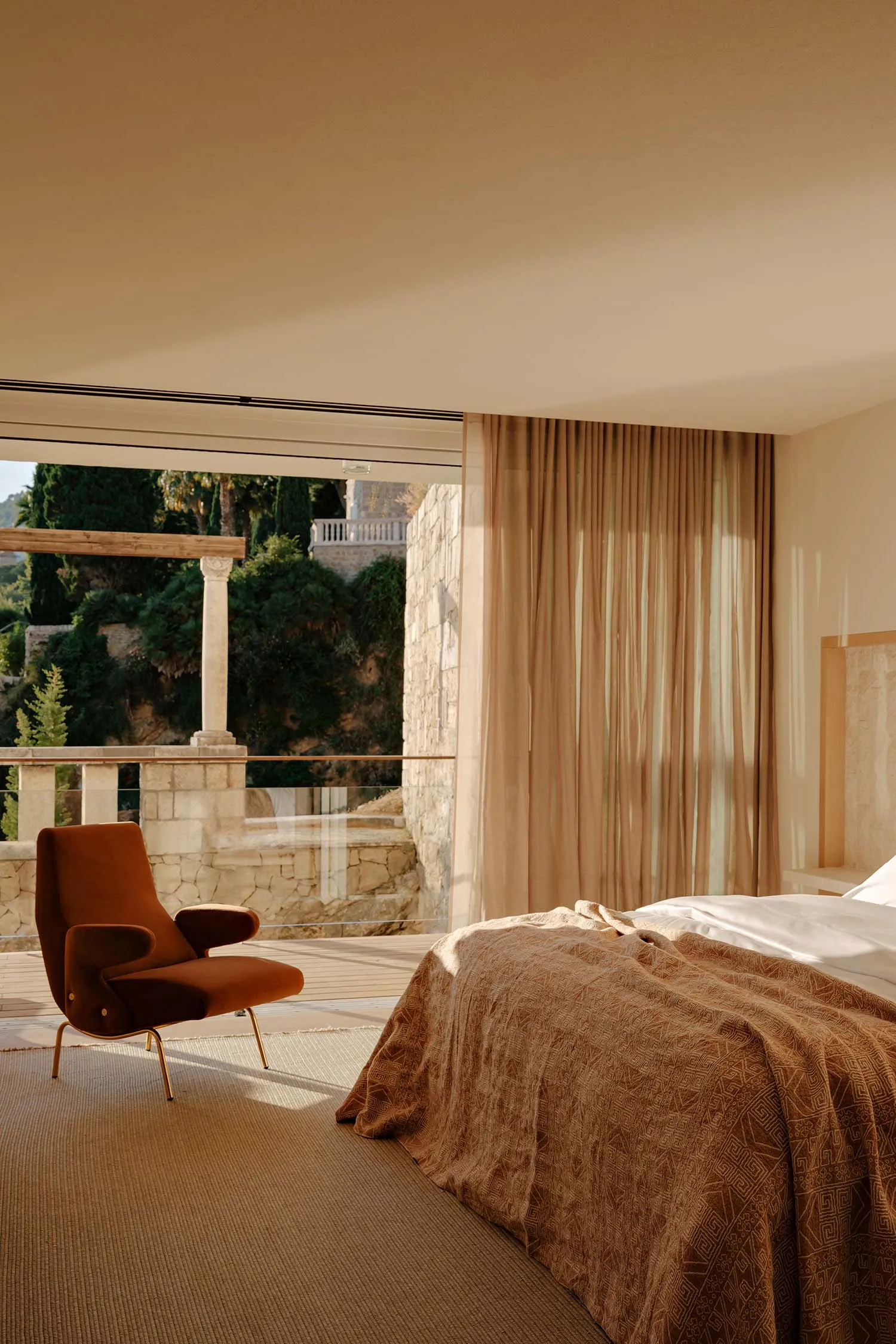 Modern Mediterranean bedroom featuring a burnt orange terracotta velvet armchair, neutral textured bedding, sheer linen curtains, and a large open terrace with ancient stone walls and lush greenery.