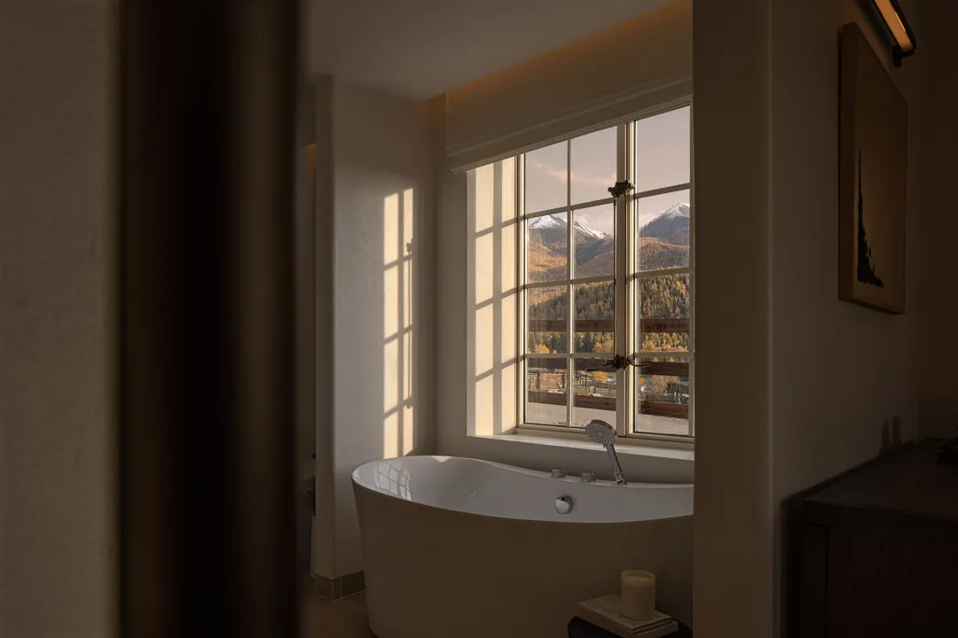 A serene bathroom featuring a white freestanding bathtub positioned in front of a large multi-pane window with a breathtaking view of snow-capped mountains and autumn forest.