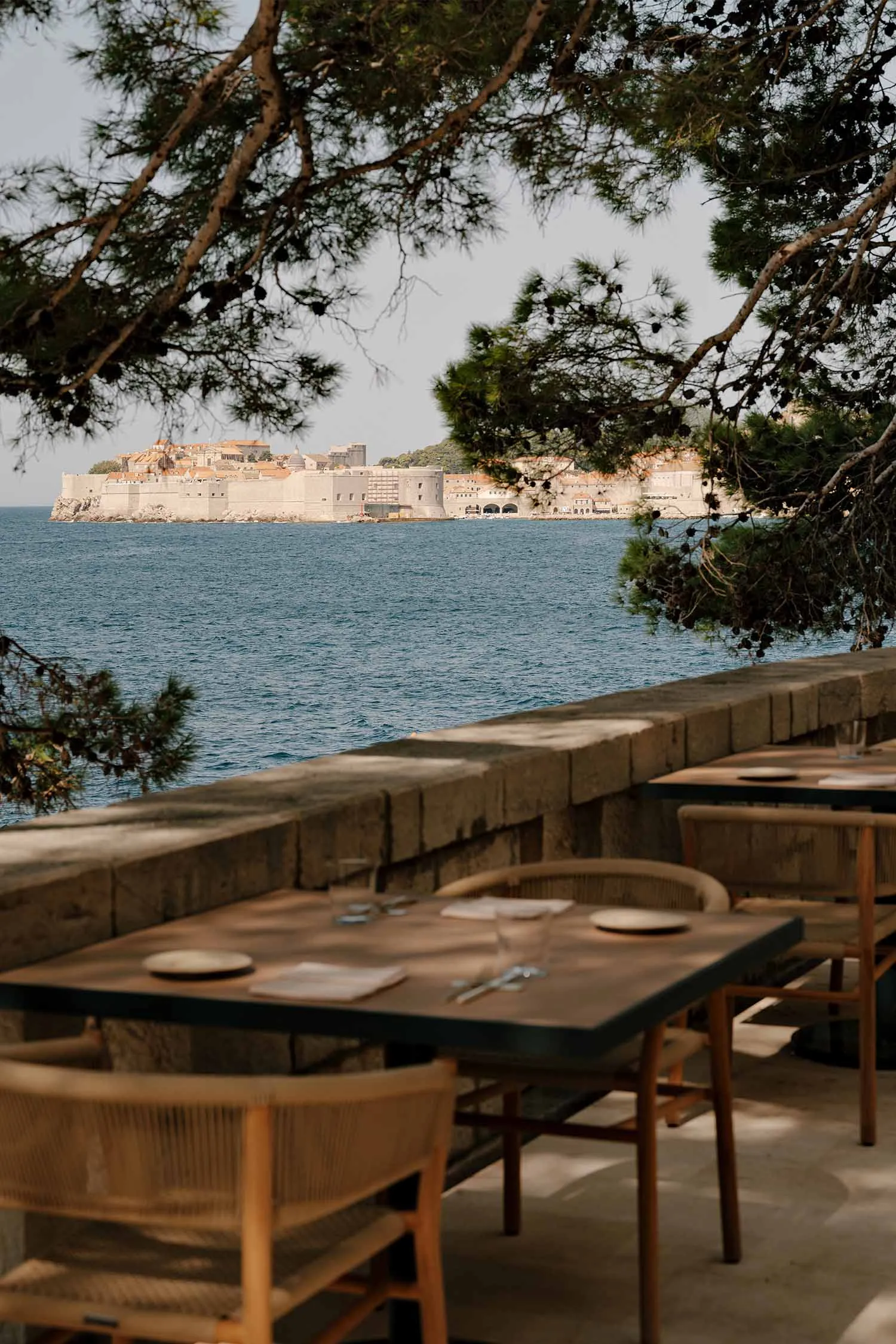Wooden dining tables and rattan chairs on a stone terrace overlooking the Adriatic Sea and the old city walls of Dubrovnik, Croatia.