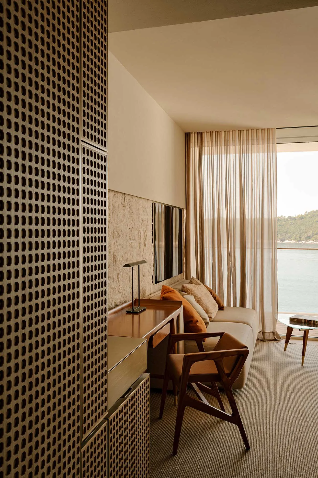 Modern coastal living room featuring a walnut console, wooden armchair, beige sofa with orange cushions, perforated wooden screen, and large window overlooking the sea.