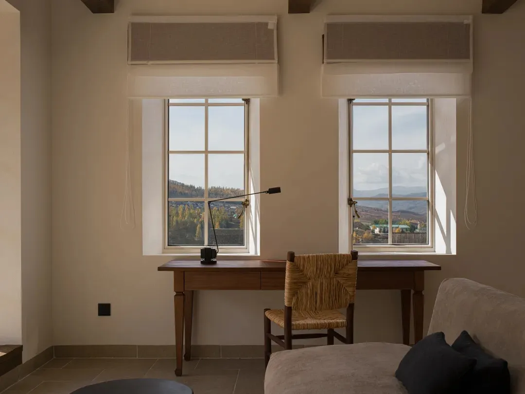 Minimal home office featuring a walnut writing desk, woven rattan chair, black articulated lamp, and two large windows overlooking forested mountains.