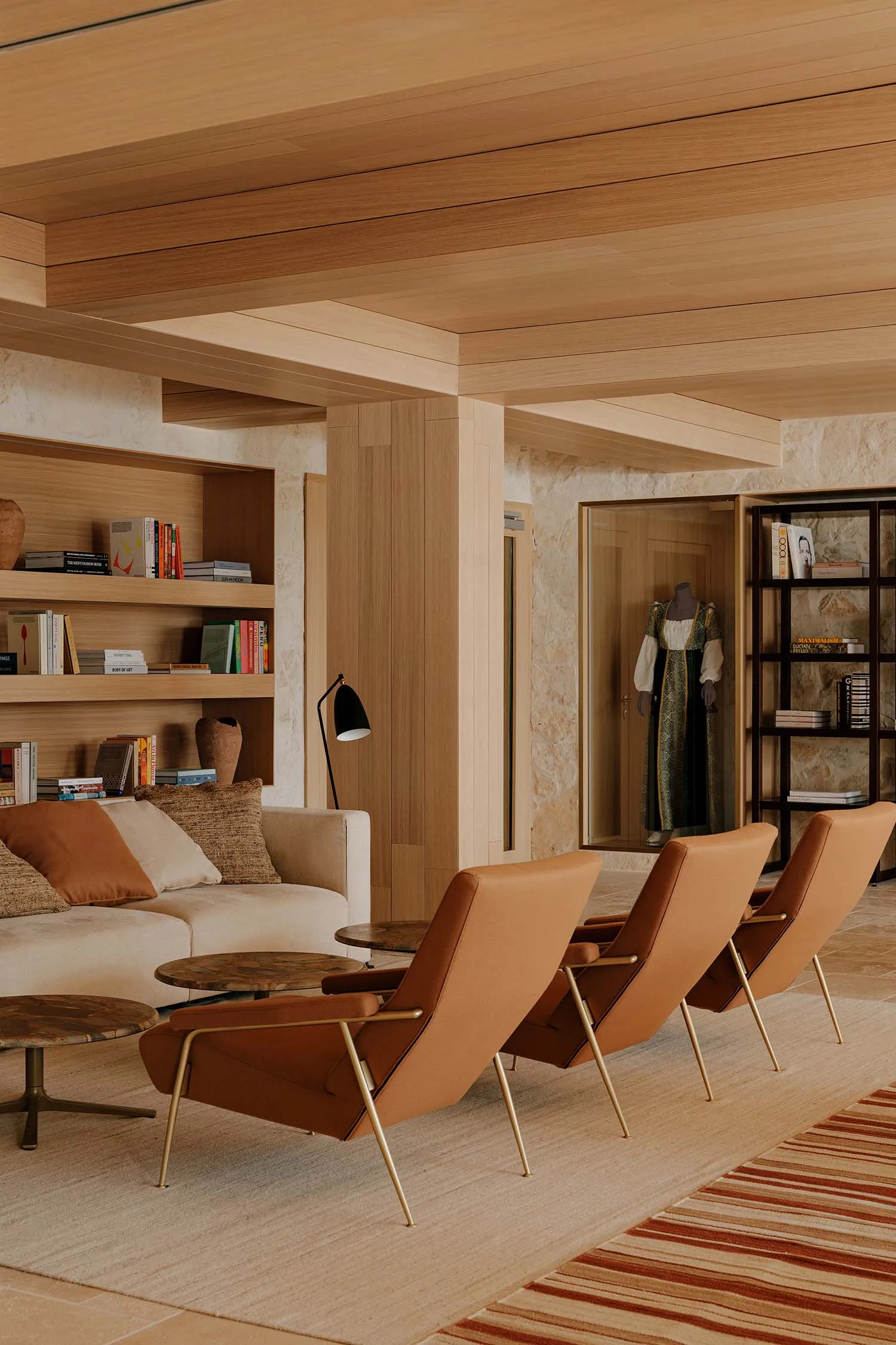 Modern minimalist living room featuring light oak wood paneling, built-in bookshelves, three cognac leather lounge chairs with brass legs, a cream sofa with terracotta cushions, and a striped area rug.