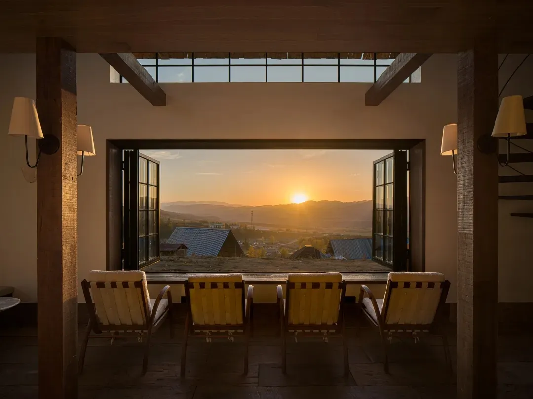 Interior view of a modern rustic sunroom with four wooden lounge chairs facing an open vista of mountains at sunrise, framed by heavy timber beams and large glass doors.