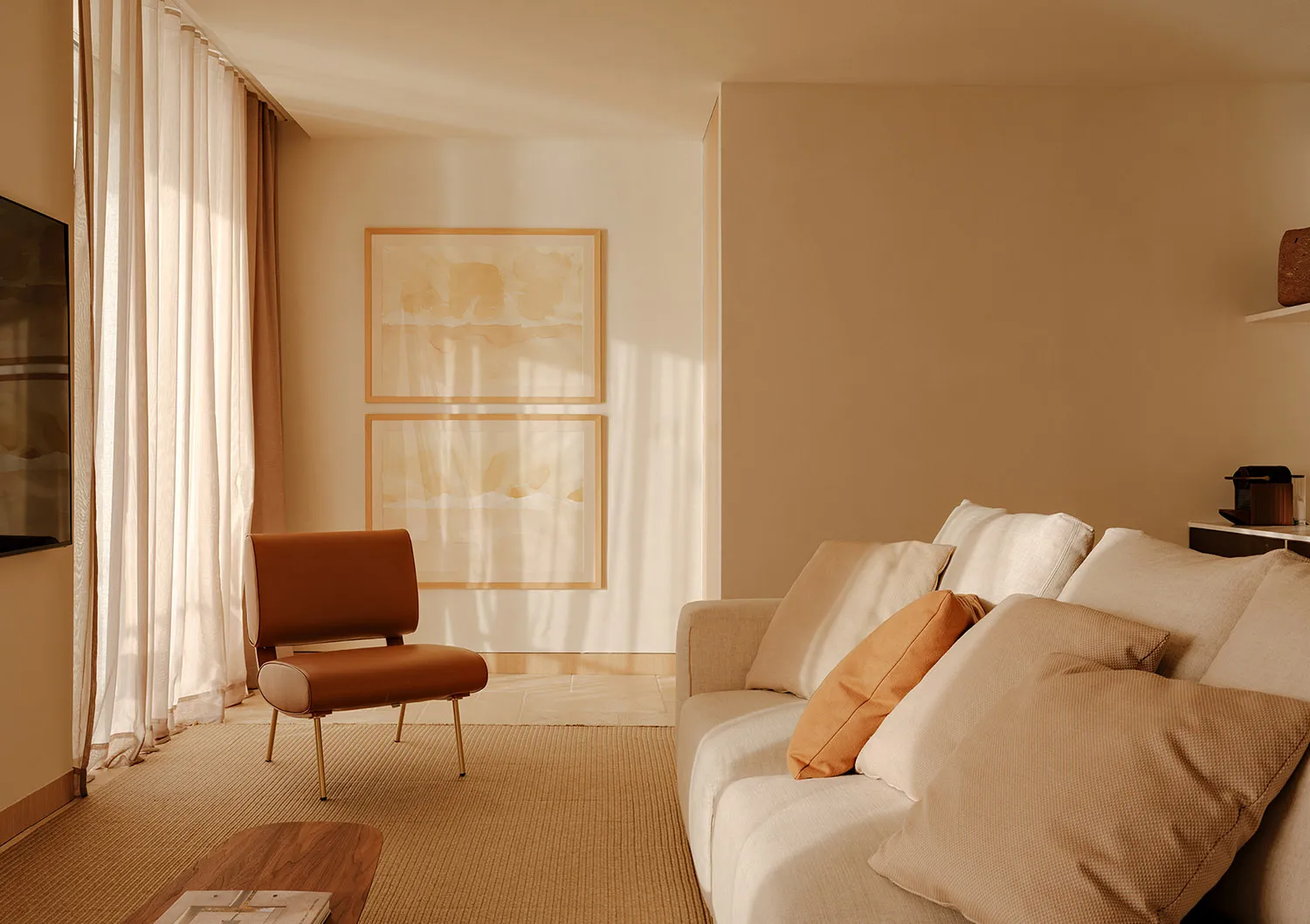 Minimalist living room featuring a cream boucle sofa with terracotta and beige cushions, a burnt orange leather accent chair with brass legs, two abstract watercolor artworks in wooden frames, sheer linen curtains, and warm natural sunlight creating soft shadows on beige walls.