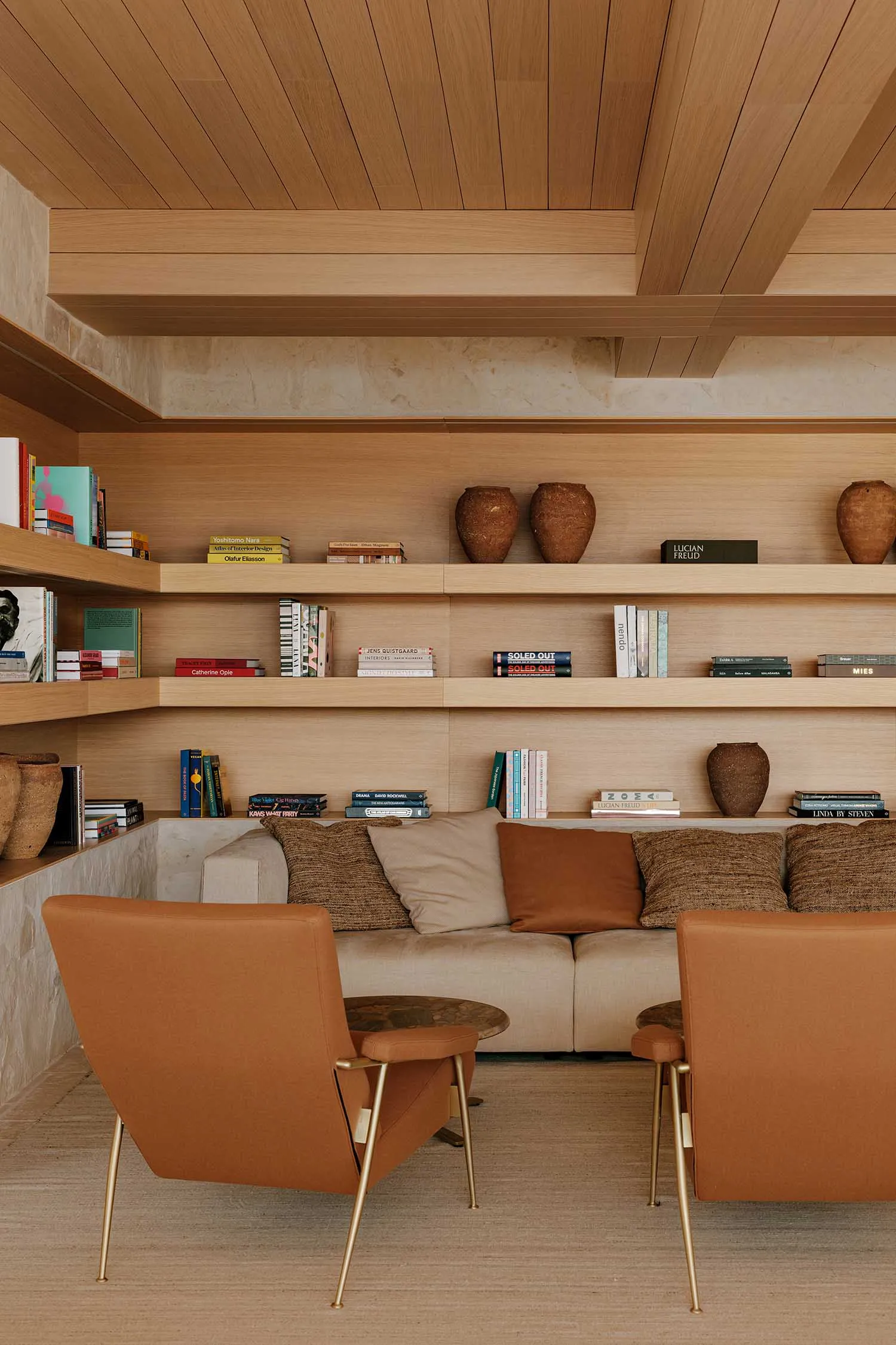 Modern living room featuring light oak built-in bookshelves, a beige sofa with terracotta and textured cushions, two burnt-orange leather armchairs with brass legs, and decorative pottery.
