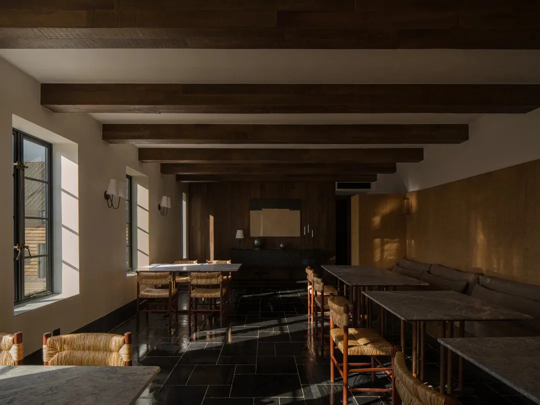 Modern rustic restaurant interior with dark wooden ceiling beams, black tiled floor, marble dining tables, and woven rattan dining chairs bathed in natural sunlight.