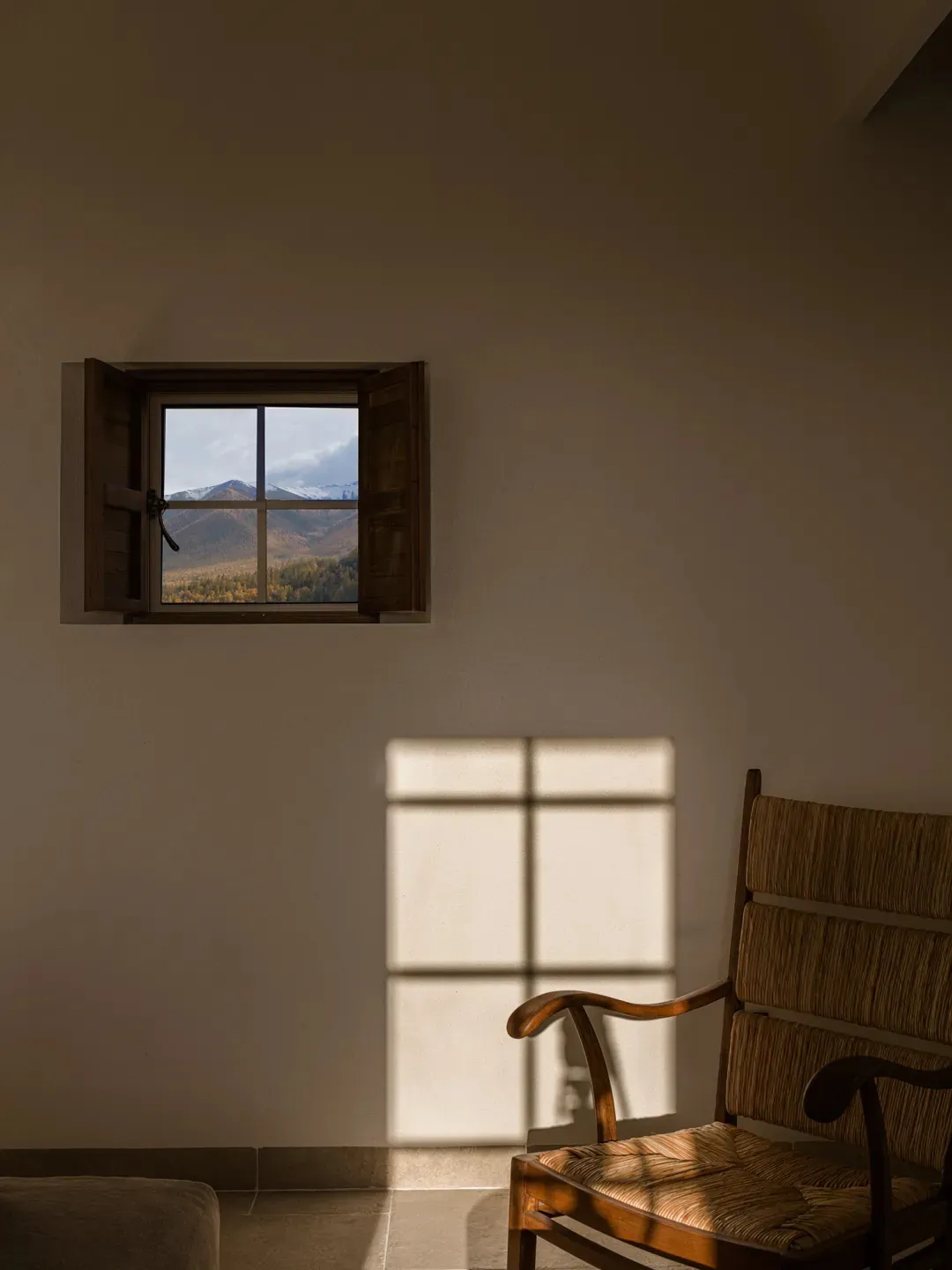 A wooden armchair with woven rattan seat and backrest placed beside a small open window revealing a scenic mountain landscape with autumn foliage and snow-capped peaks.