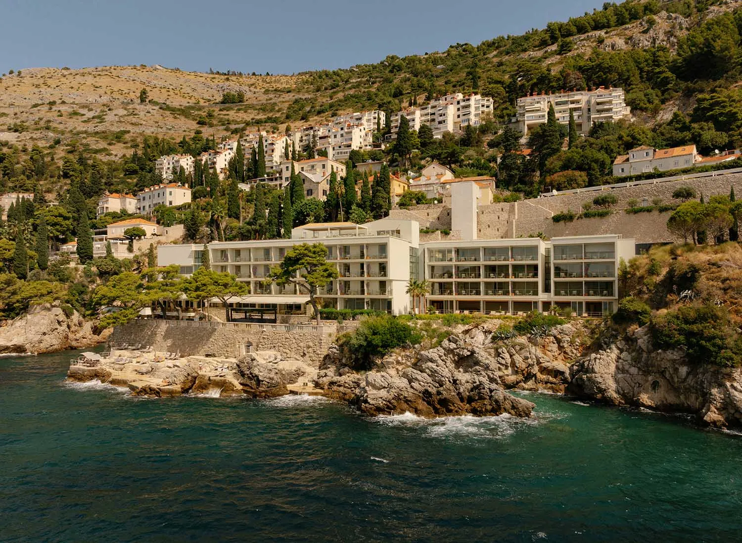 A contemporary white hotel with extensive glass windows built directly on rocky cliffs above the deep blue sea, with a hillside town in the background under a clear sky.
