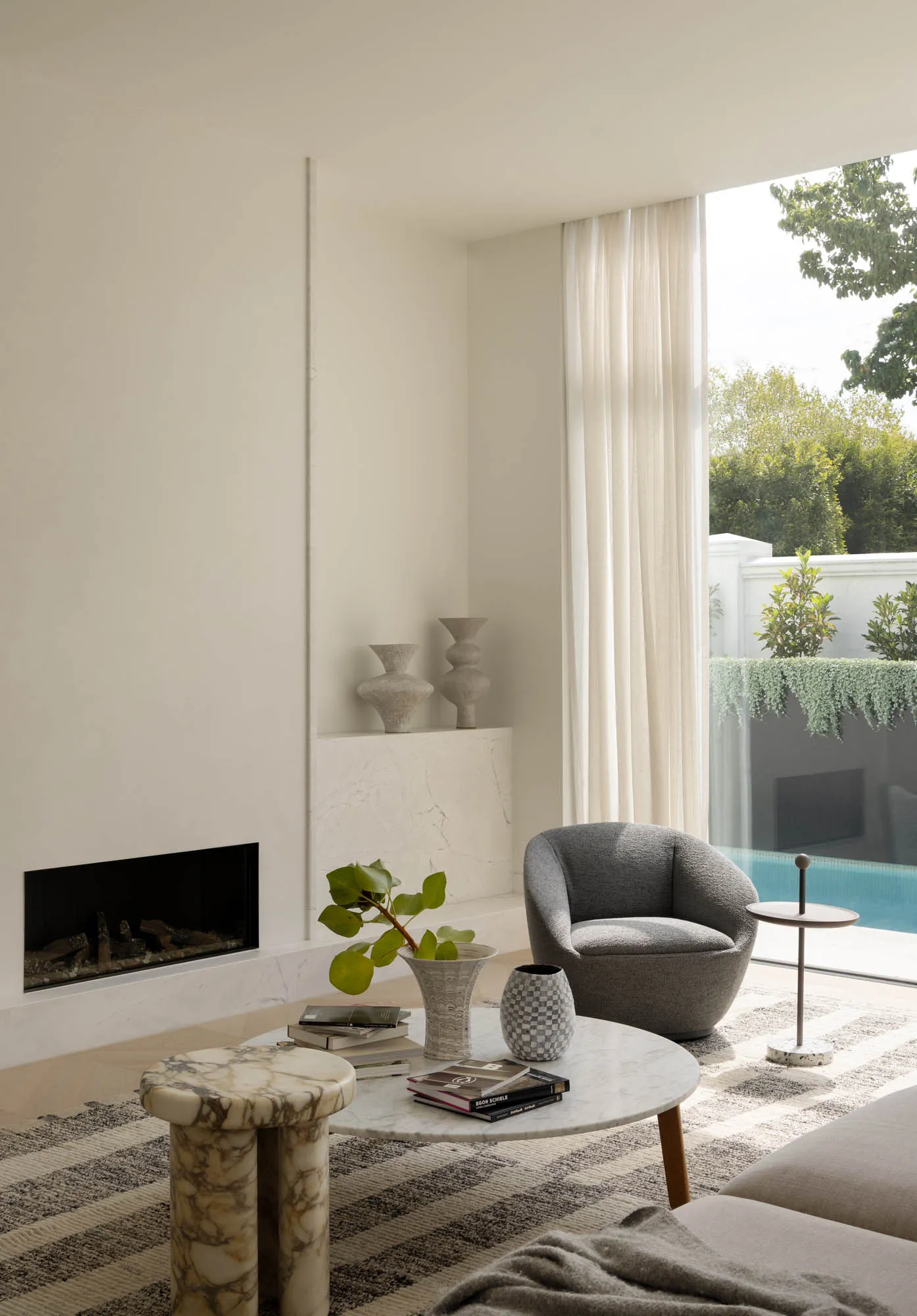 Minimalist living room featuring a marble fireplace, curved grey bouclé armchair, round marble coffee table, and large glass doors opening to a swimming pool and garden.