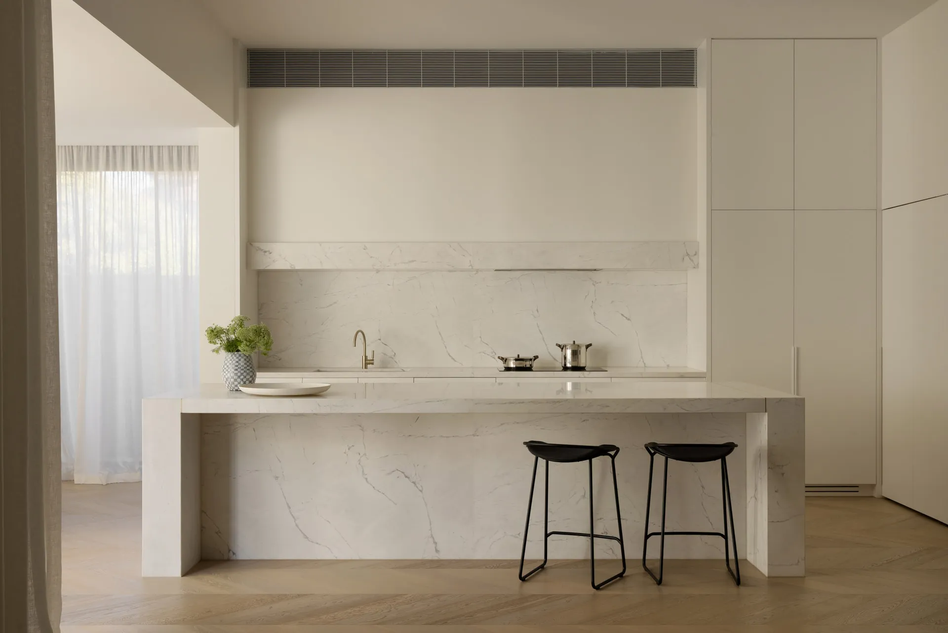 Modern minimalist kitchen featuring a large white marble island with two black wireframe stools, matching marble backsplash and counter, brass faucet, and soft natural light from a window with sheer curtains.