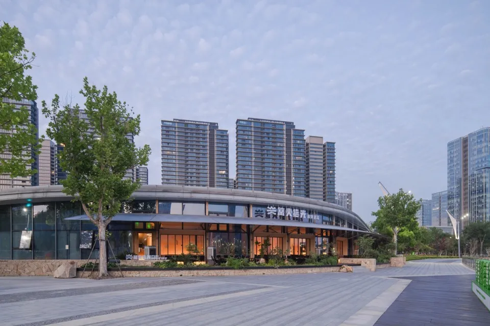 現代弧形玻璃與混凝土社區中心,內部燈光亮起,位於寬敞都市廣場,前有樹木,背景為高層住宅大樓,拍攝於黃昏時分。