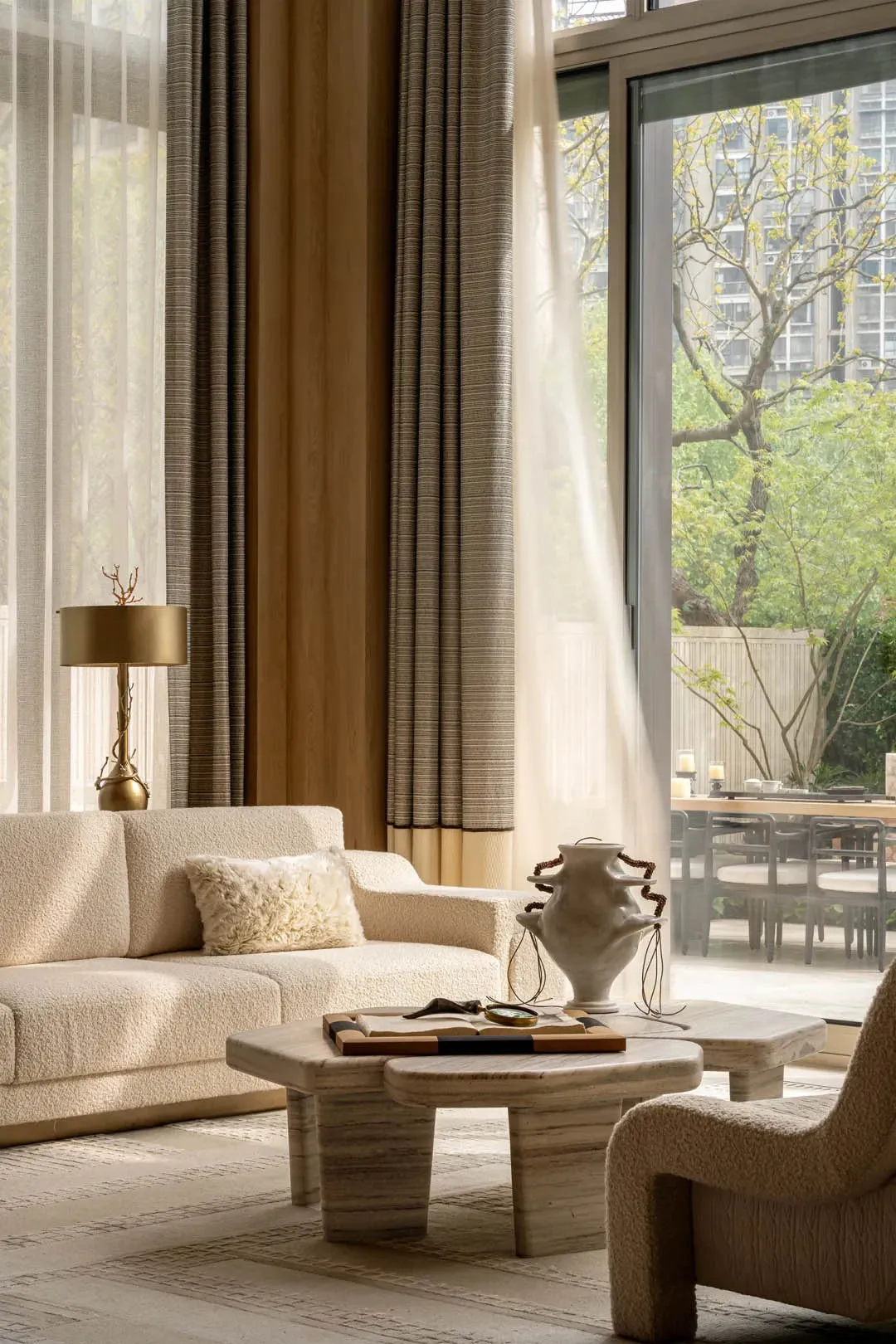 Modern living room featuring a cream boucle sofa, travertine coffee tables, sculptural ceramic vase, and large glass doors opening to a green garden view.