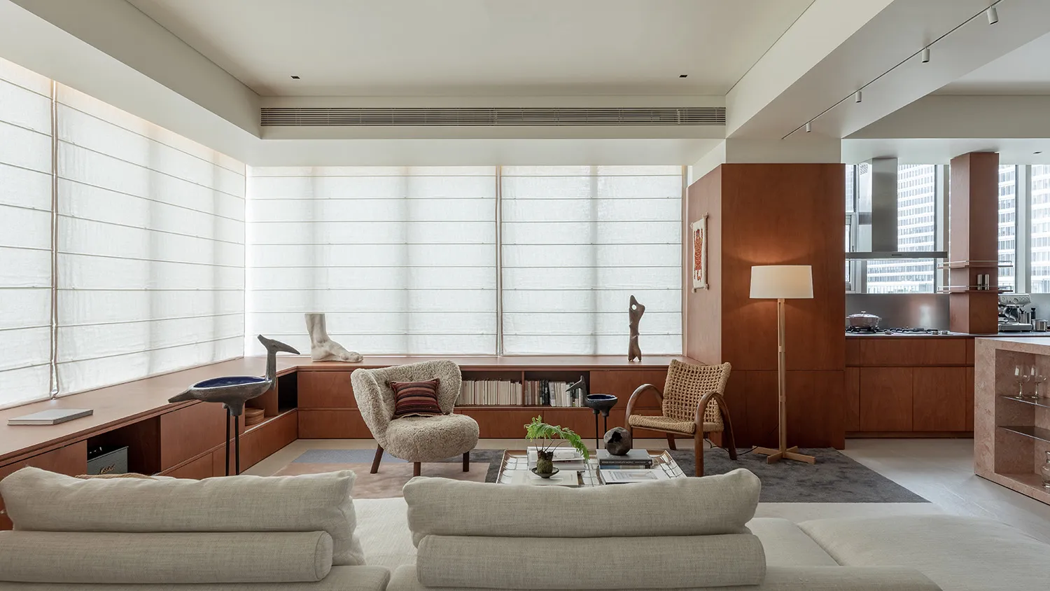 Modern minimalist living room featuring extensive walnut built-in cabinetry, a large boucle armchair, a rattan lounge chair, and a light-colored sectional sofa with large windows covered by sheer roman shades.