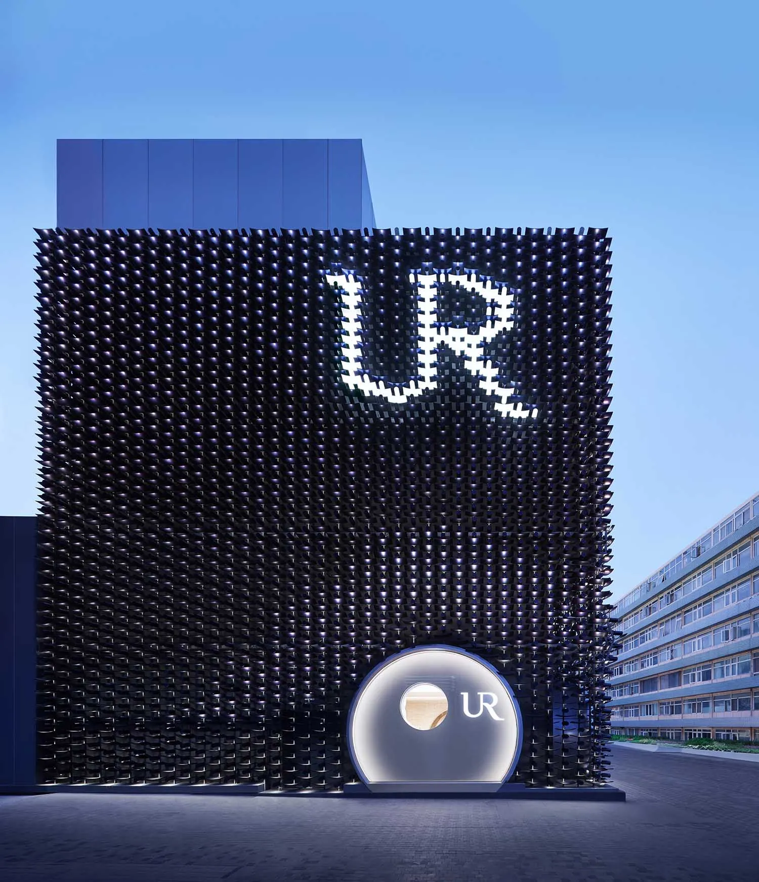 夜晚的現代建築,黑色穿孔金屬立面帶有大型發光 UR 標誌與圓形入口結構。