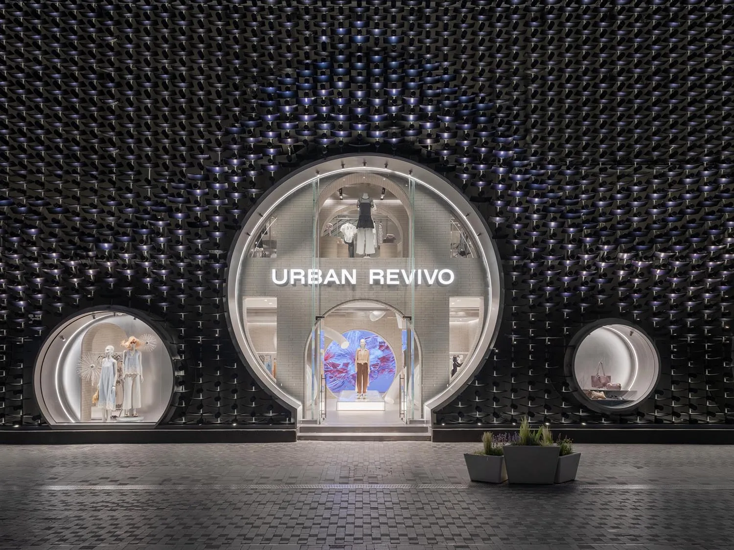 Night view of Urban Revivo fashion store featuring a large circular glass entrance set in a dramatic black textured sculptural wall with smaller circular display windows on either side.