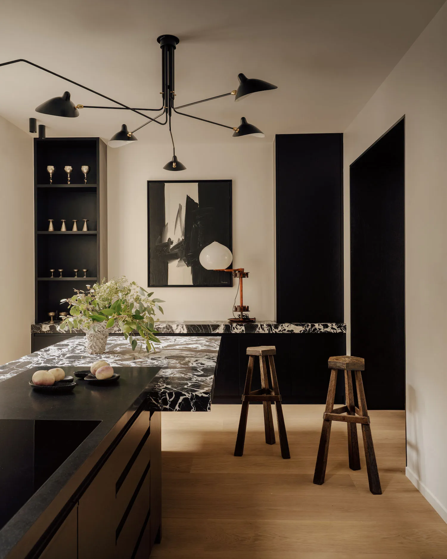 Modern kitchen with black marble island, dark cabinetry, two rustic wooden bar stools, large black chandelier, and abstract black and white artwork on the wall.