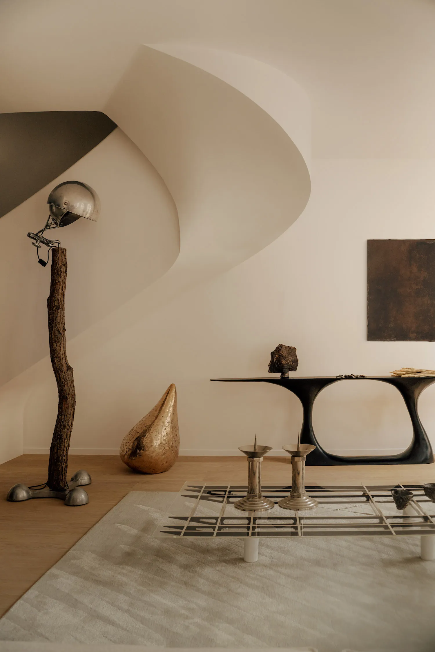Modern minimalist living room featuring a sculptural black console table, a floor lamp made from a real tree trunk with a vintage helmet shade, a textured gold teardrop sculpture, and a glass coffee table with metallic candle holders on a neutral rug.