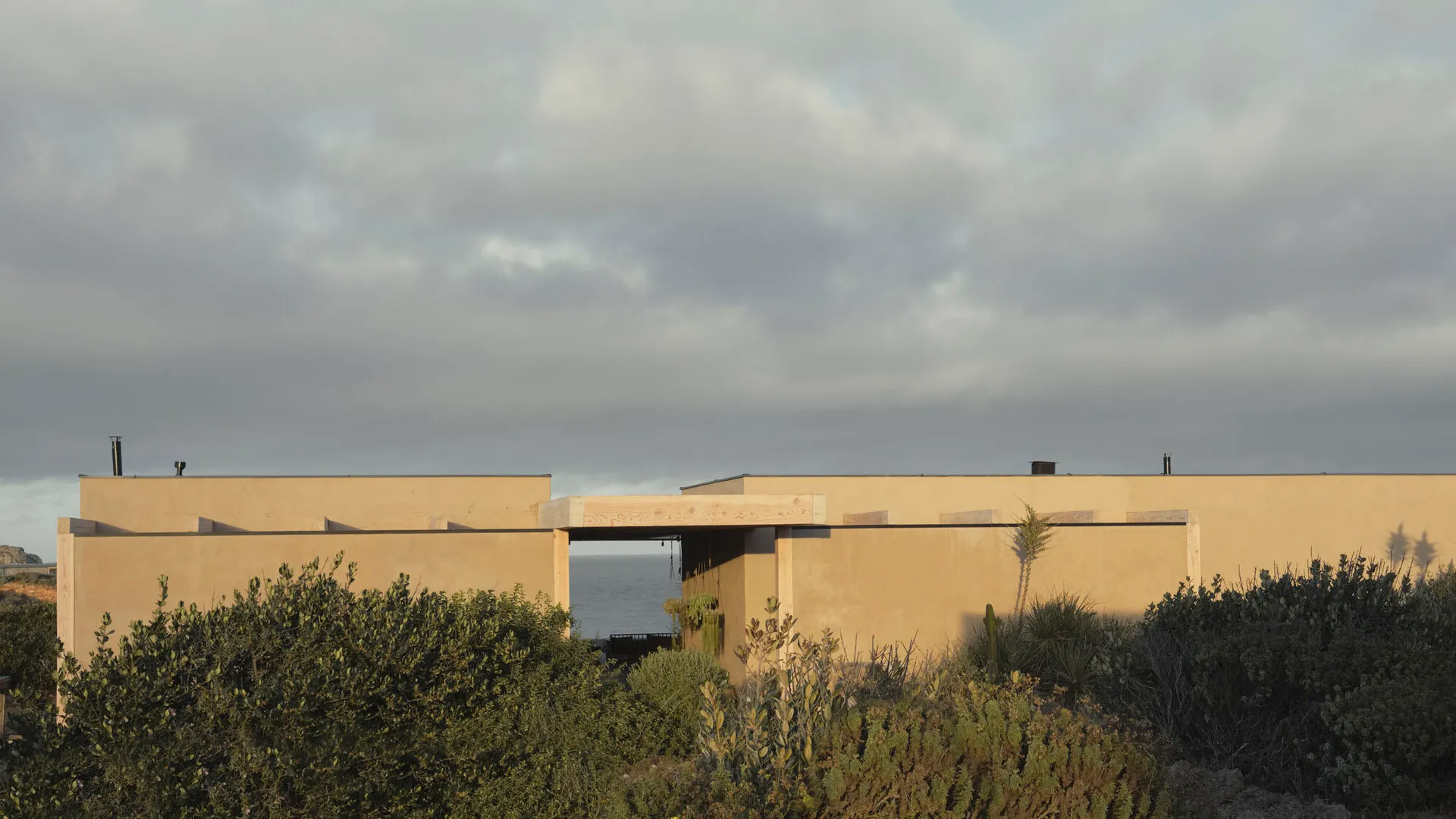 Modern single-story stucco house with wooden beams and a central courtyard opening that perfectly frames the blue ocean horizon, surrounded by coastal native shrubs under a cloudy sky.