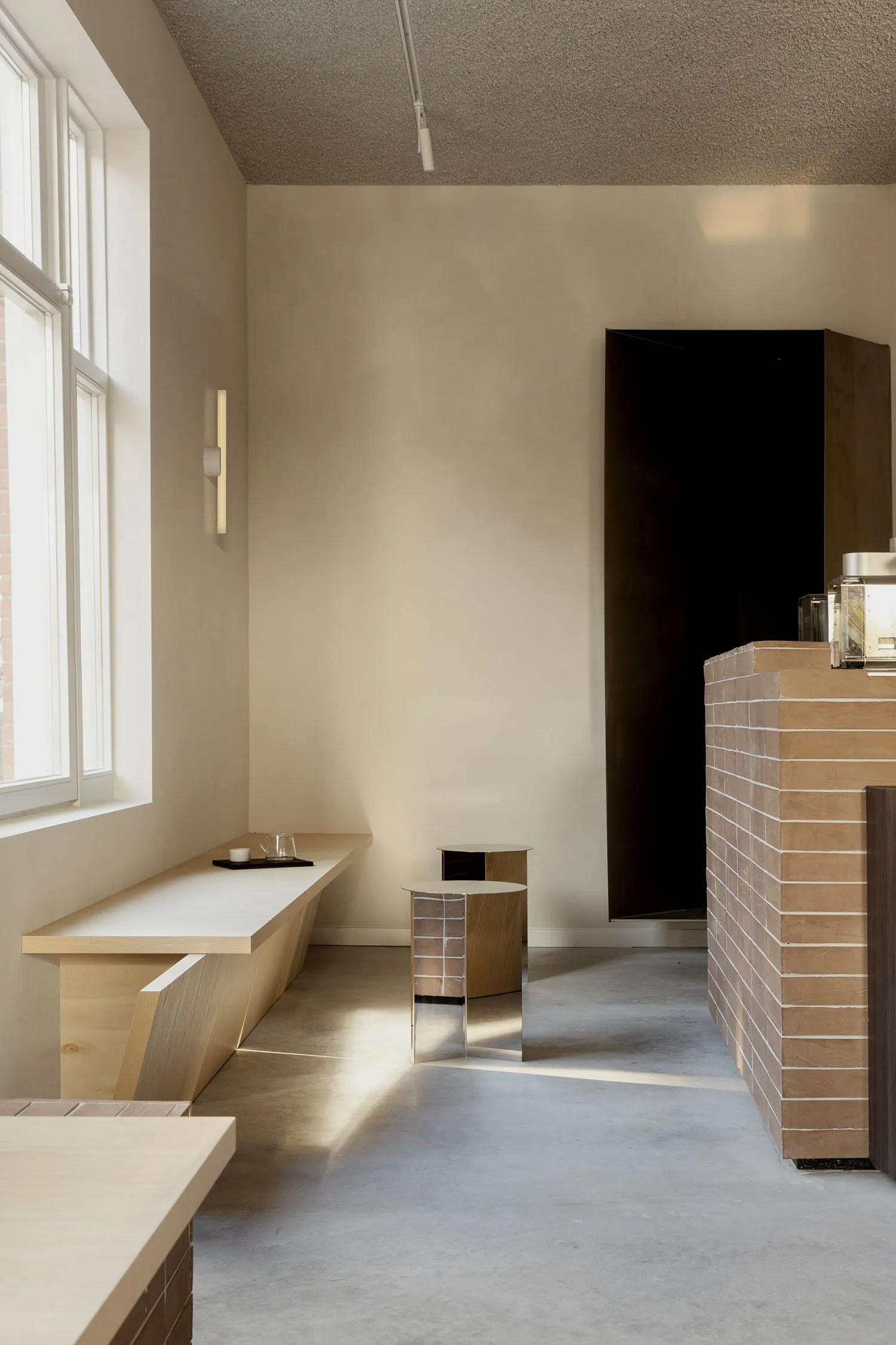 Minimalist cafe interior featuring a long light wood bench under a window, cylindrical wooden side tables with metallic accents, and a terracotta brick counter against a neutral beige wall.