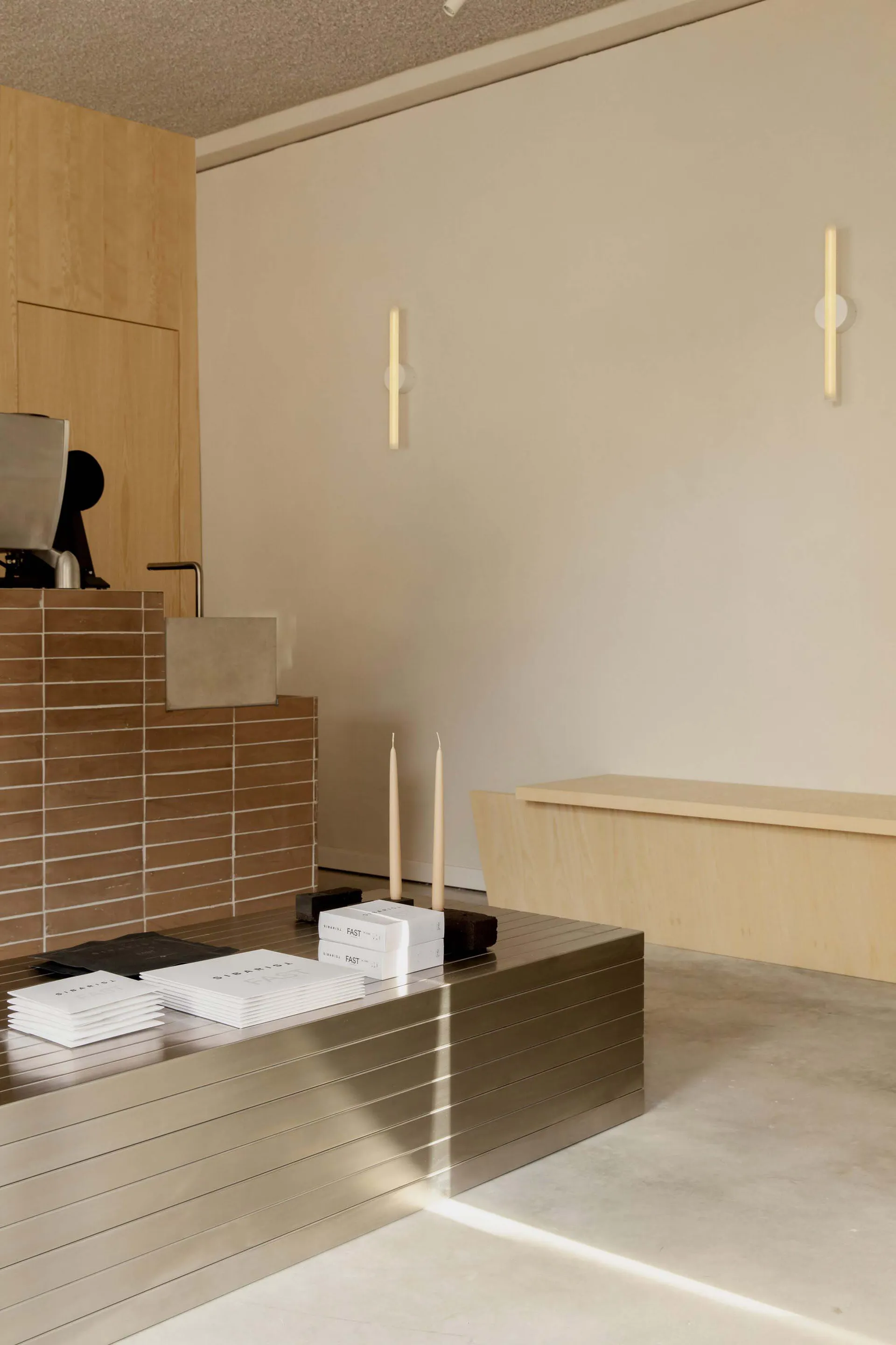Modern minimalist reception desk made of horizontal stainless steel slats in the foreground, with a brown tiled counter and light wood cabinetry in the background, two tall candles and stacked brochures on the desk.