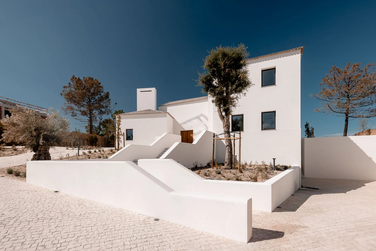 Modern white stucco Mediterranean villa with stepped white walls, terraced garden, olive trees, and bright blue sky in a sunny coastal setting.