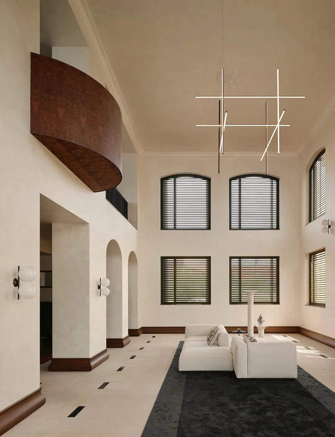 Spacious double-height living room featuring a curved walnut balcony, white L-shaped sectional sofa on a dark rug, modern geometric chandelier, and arched doorways with dark wood trim.