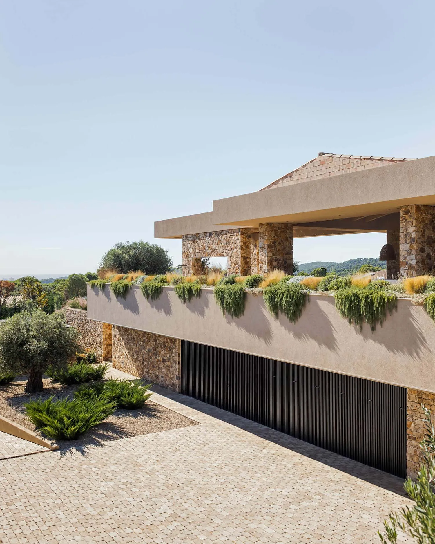 Modern Mediterranean villa exterior with stone walls, concrete overhang, cascading rooftop garden, and paved courtyard under bright sunlight.