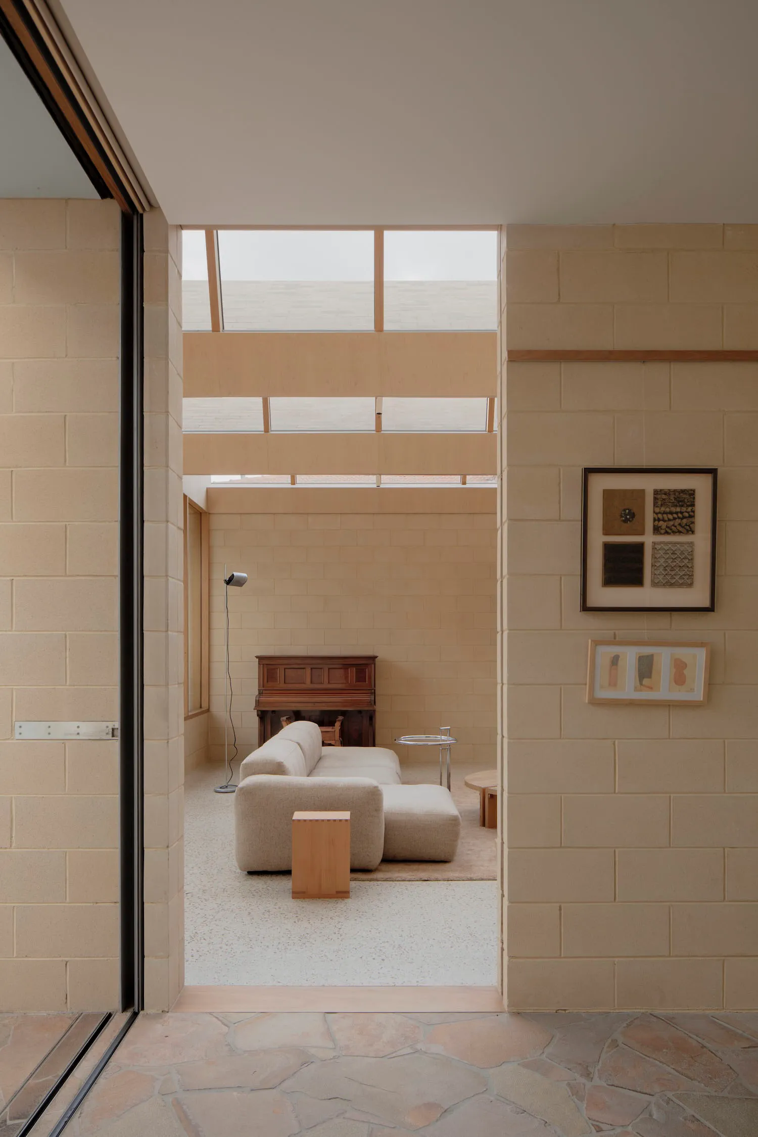 View into a minimalist living room featuring light-colored concrete block walls, a large modular beige sofa on terrazzo flooring, wooden accents, and high clerestory windows allowing soft natural light.