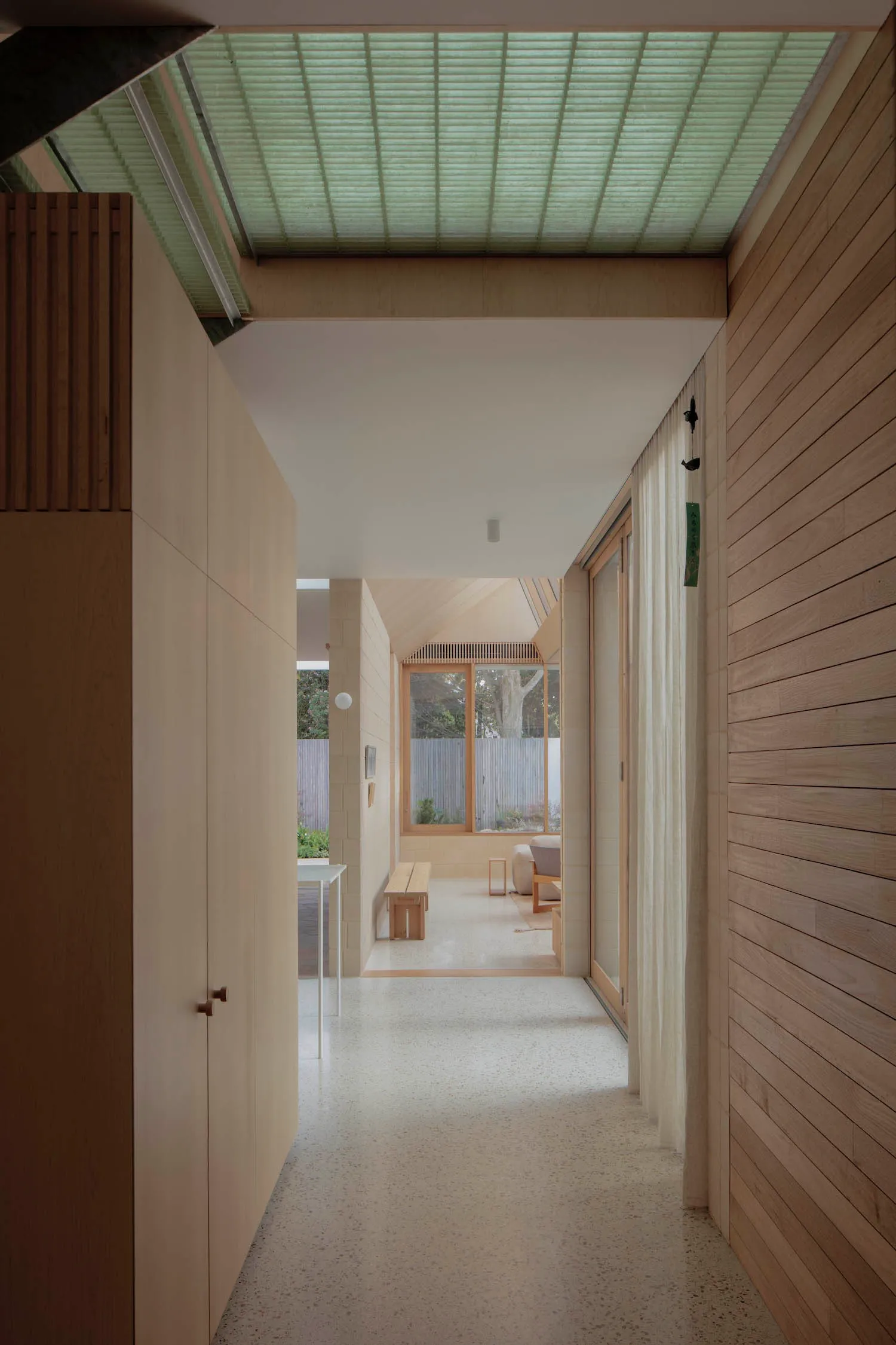 Long modern wooden corridor with light wood paneling, polished terrazzo floor, large clerestory skylight with blinds, and view to a bright garden courtyard.