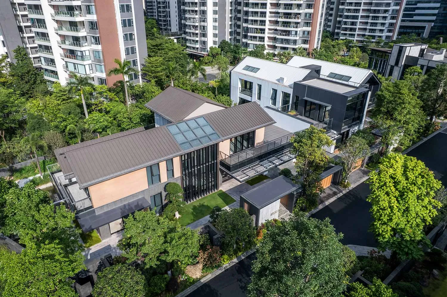 Aerial view of a contemporary two-story villa featuring large glass windows, dark metal roof, warm beige walls, and surrounded by dense green trees and a manicured garden in a suburban setting with apartment buildings in the background.