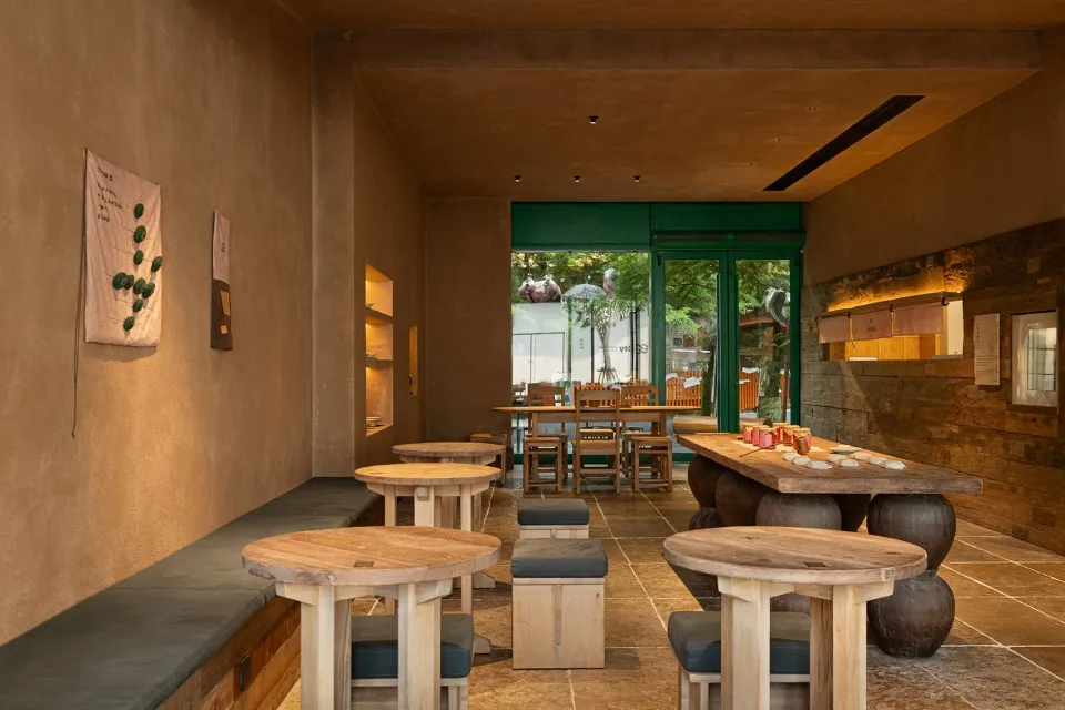Modern wabi-sabi style restaurant interior with round solid wood tables, concrete walls, wooden stools with dark cushions, and large green-framed glass doors opening to a courtyard.