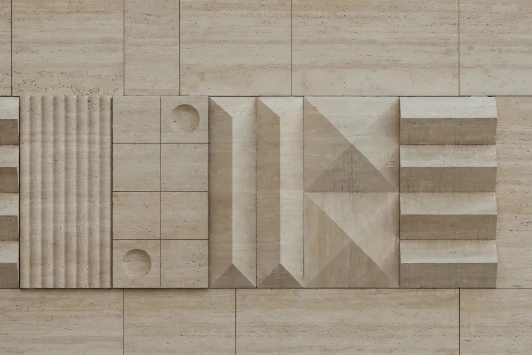 Close-up of a beige travertine stone wall featuring various three-dimensional geometric patterns including fluted columns, circular cutouts, angular pyramids, and stepped reliefs.