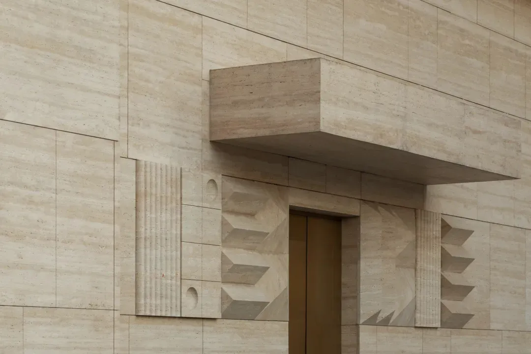 Close-up of a luxurious travertine stone building entrance featuring geometric relief patterns, fluted columns, and a cantilevered overhang above a bronze door.