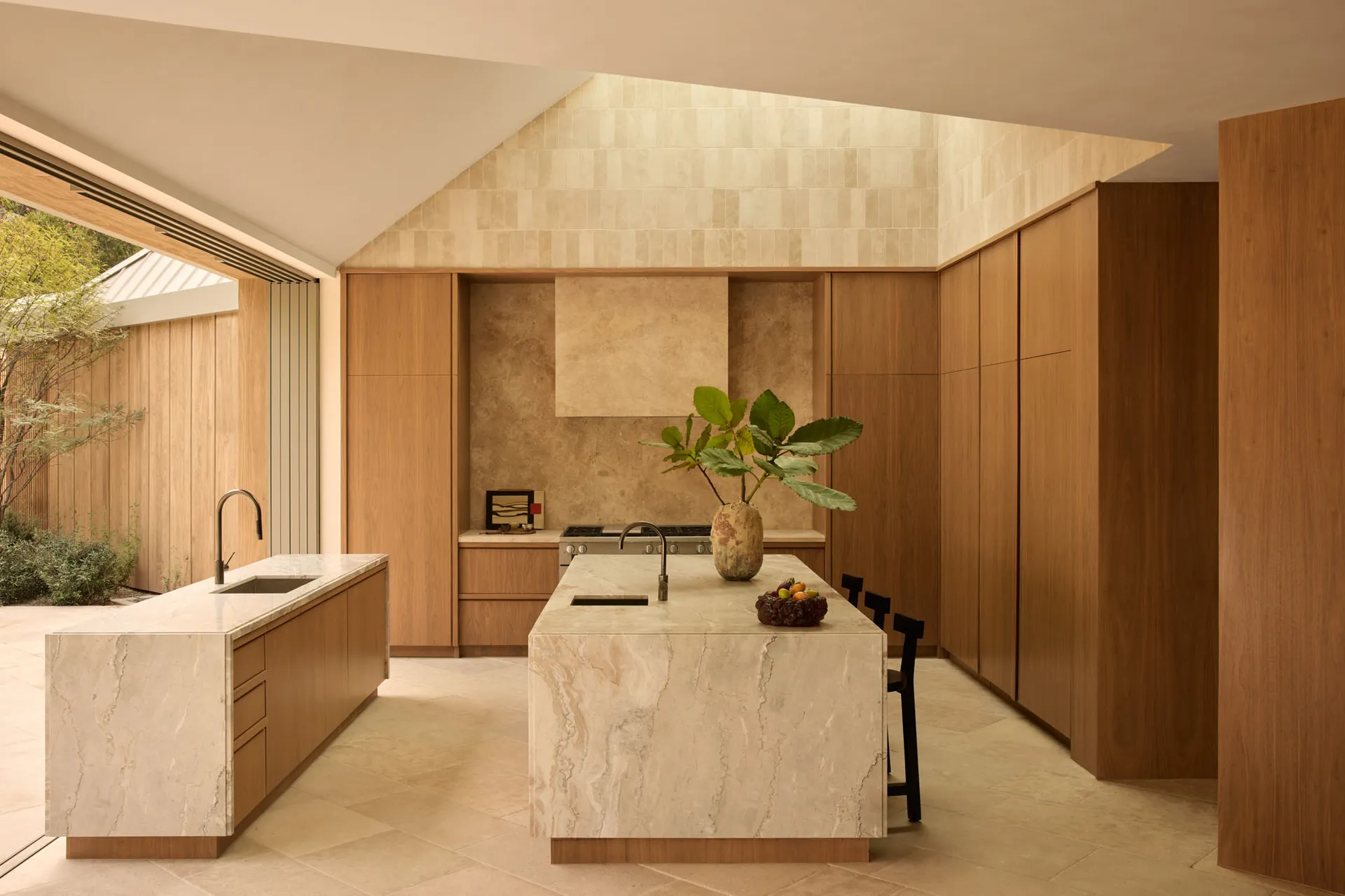 Modern kitchen featuring a large central island made of veined travertine, warm oak cabinetry, black metal chairs, and a tall skylight with textured stone walls.