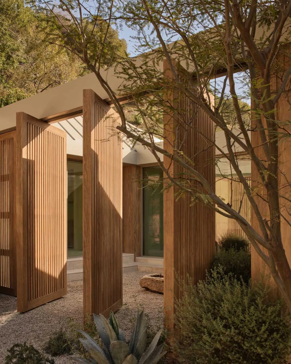 Modern outdoor courtyard featuring vertical teak wood slatted walls and doors, gravel ground, native desert plants, and a mountain backdrop under a clear blue sky.