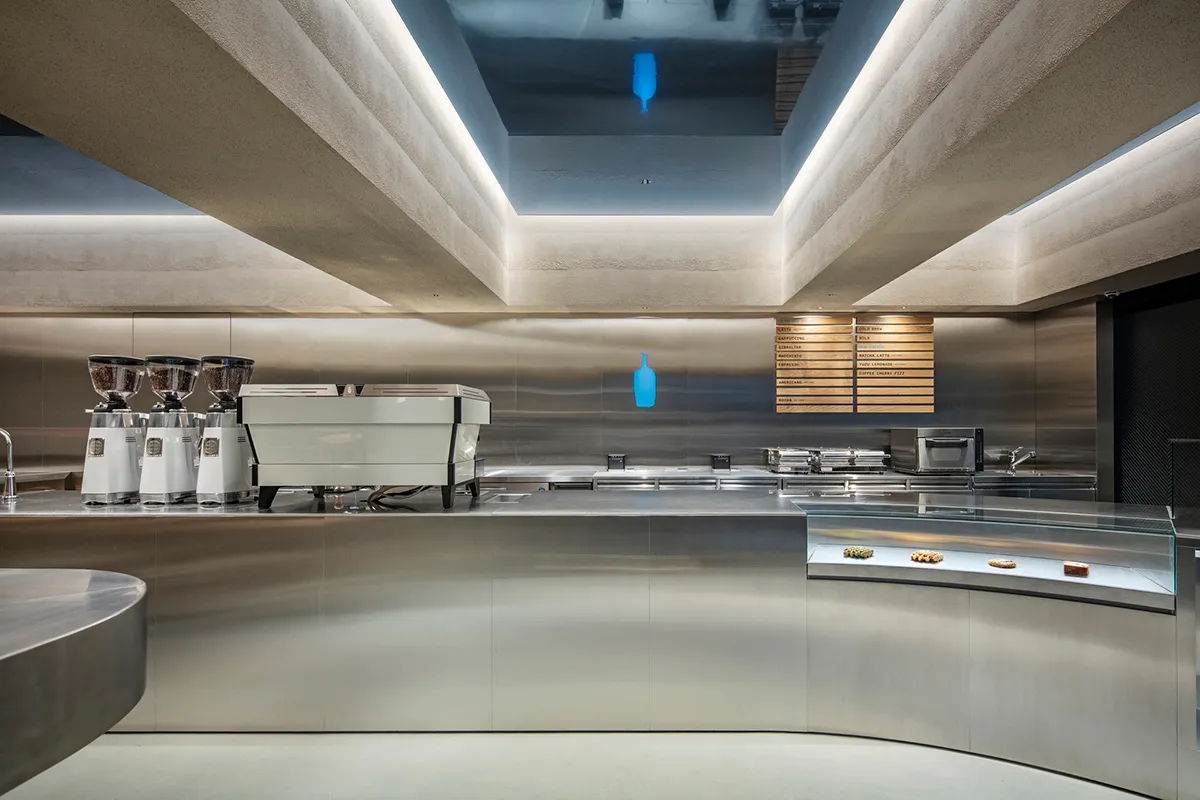 Modern minimalist coffee shop service counter made of curved stainless steel with concrete ceiling and integrated LED lighting