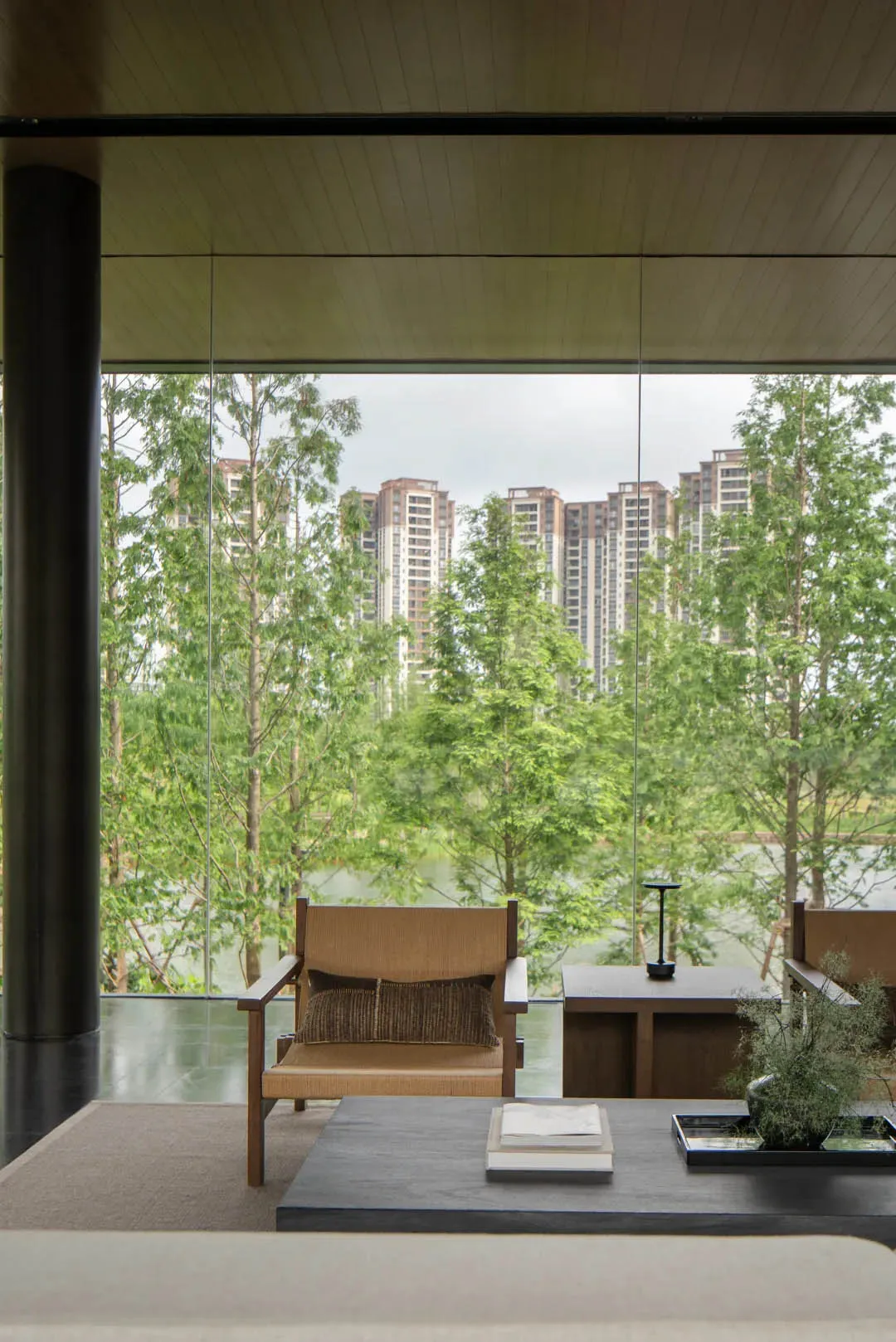 Modern minimalist living room featuring wooden armchairs, dark stone coffee table, and expansive glass windows revealing a green lakeside view with city buildings in the background.