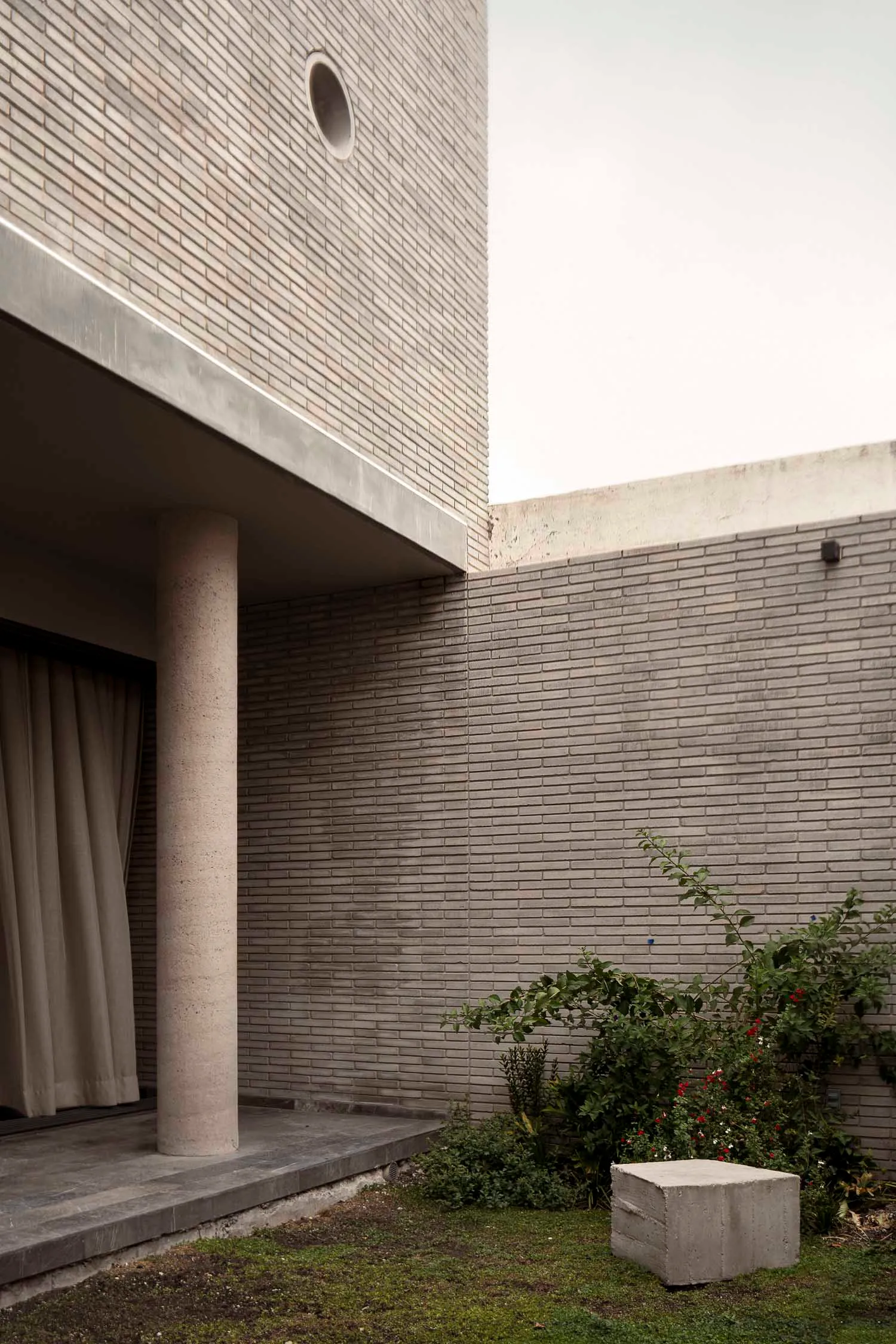 Exterior view of a modern brutalist courtyard featuring an exposed concrete column, grey brick walls, a circular window, and lush greenery with a concrete block stool.