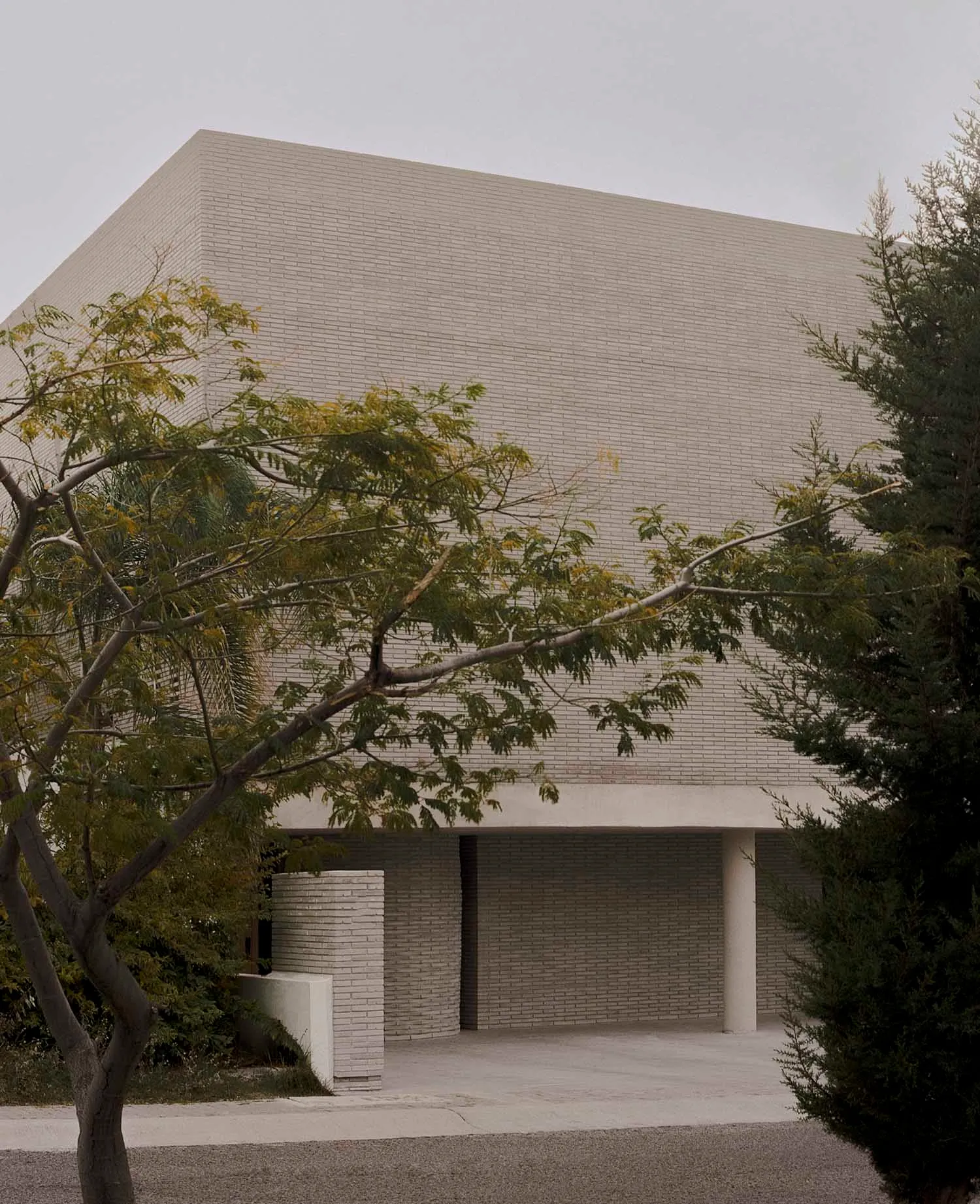 Exterior view of a brutalist concrete building with a strong geometric form, overhanging canopy, and surrounding trees under an overcast sky.